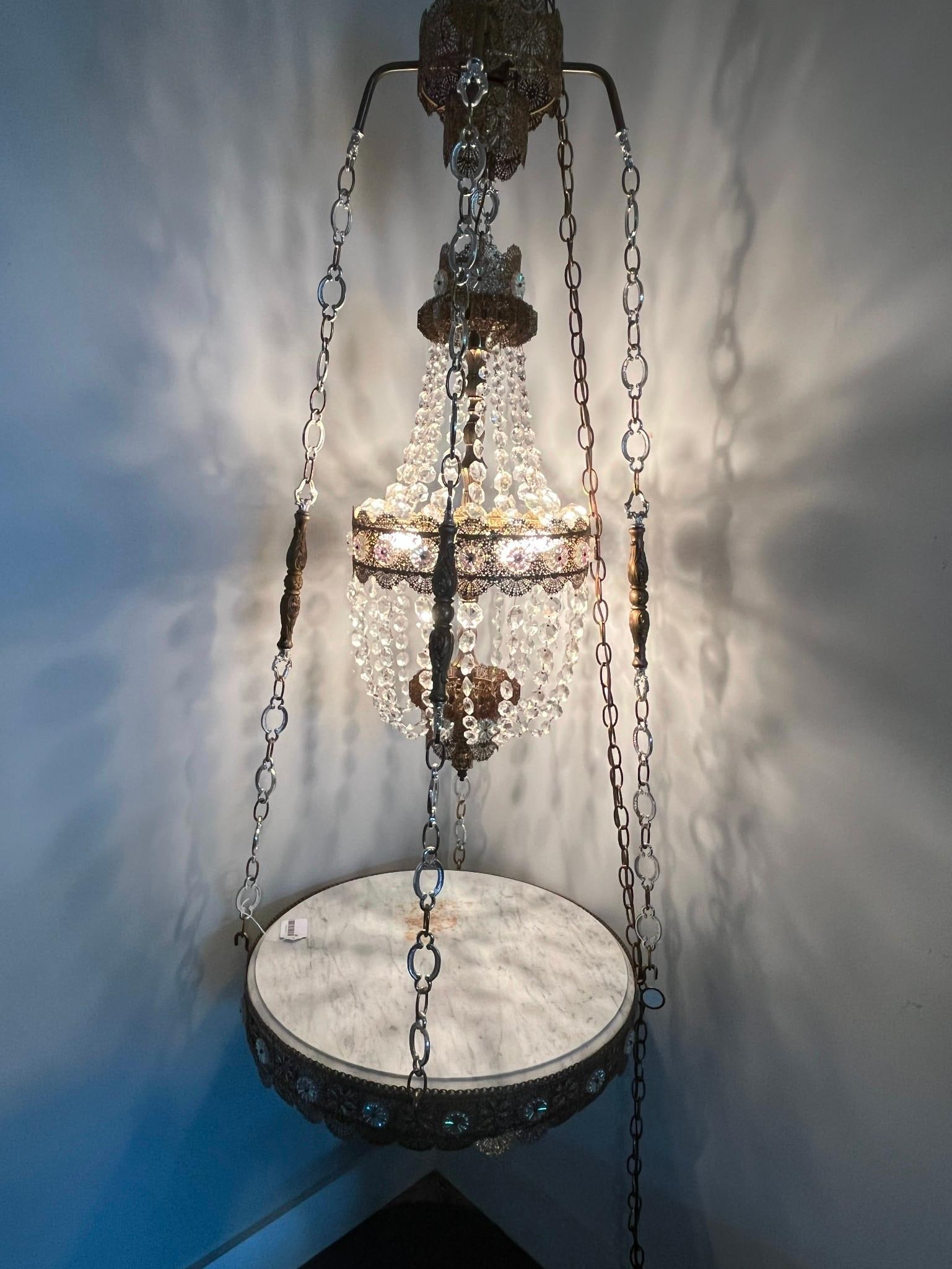 Hollywood Regency Chandelier w/ Table: A tiered Hollywood Regency style decorative hanging fixture comprising a central chandelier suspended above a circular marble tabletop. The unit is constructed from pierced metal filigree with a