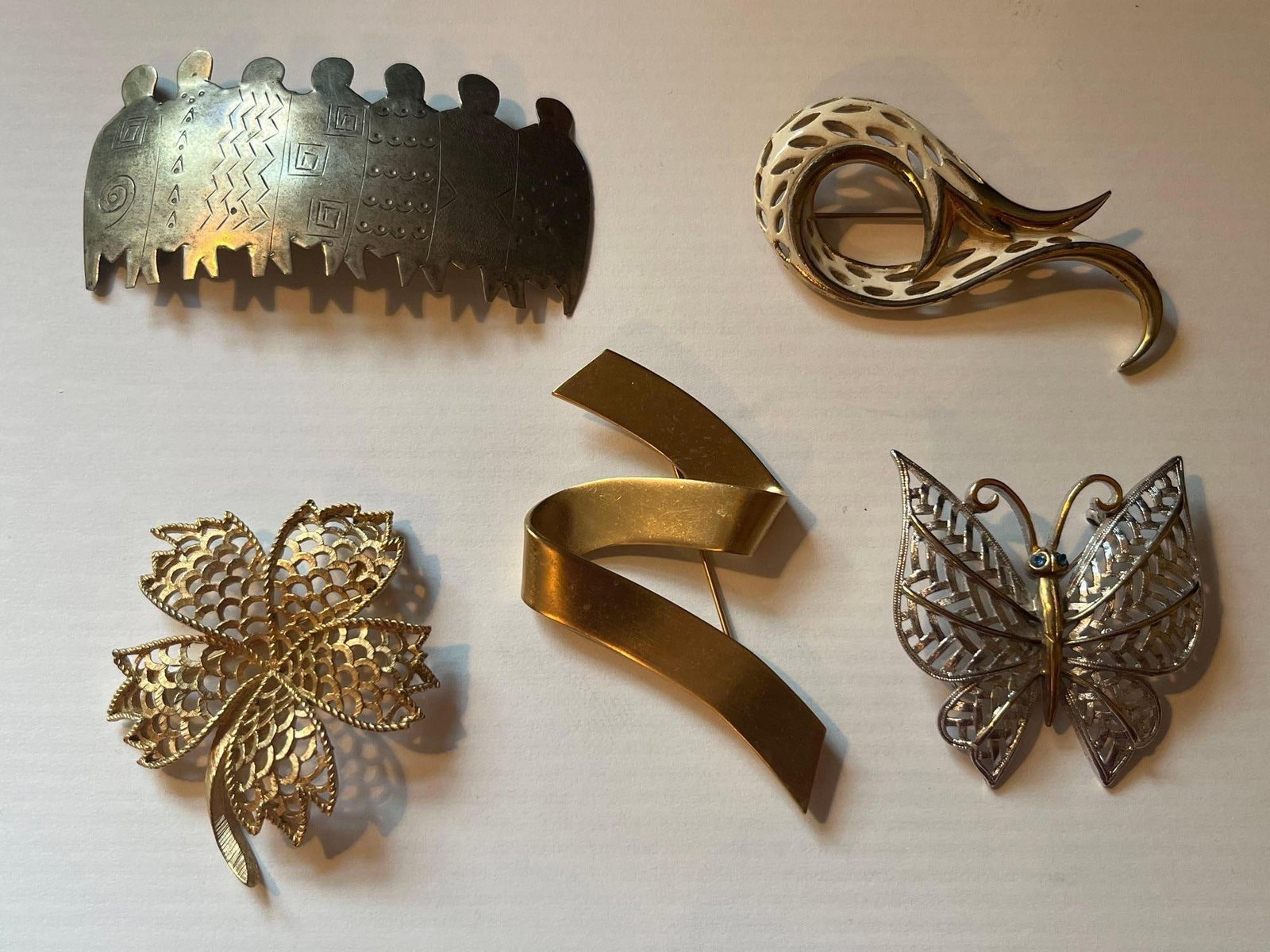 Trifari, Monet & Emmons Brooches: This collection consists of several costume jewelry brooches in various figurative and modernist styles. One rectangular brooch in silver-toned base metal features seven stylized humanoid figures with