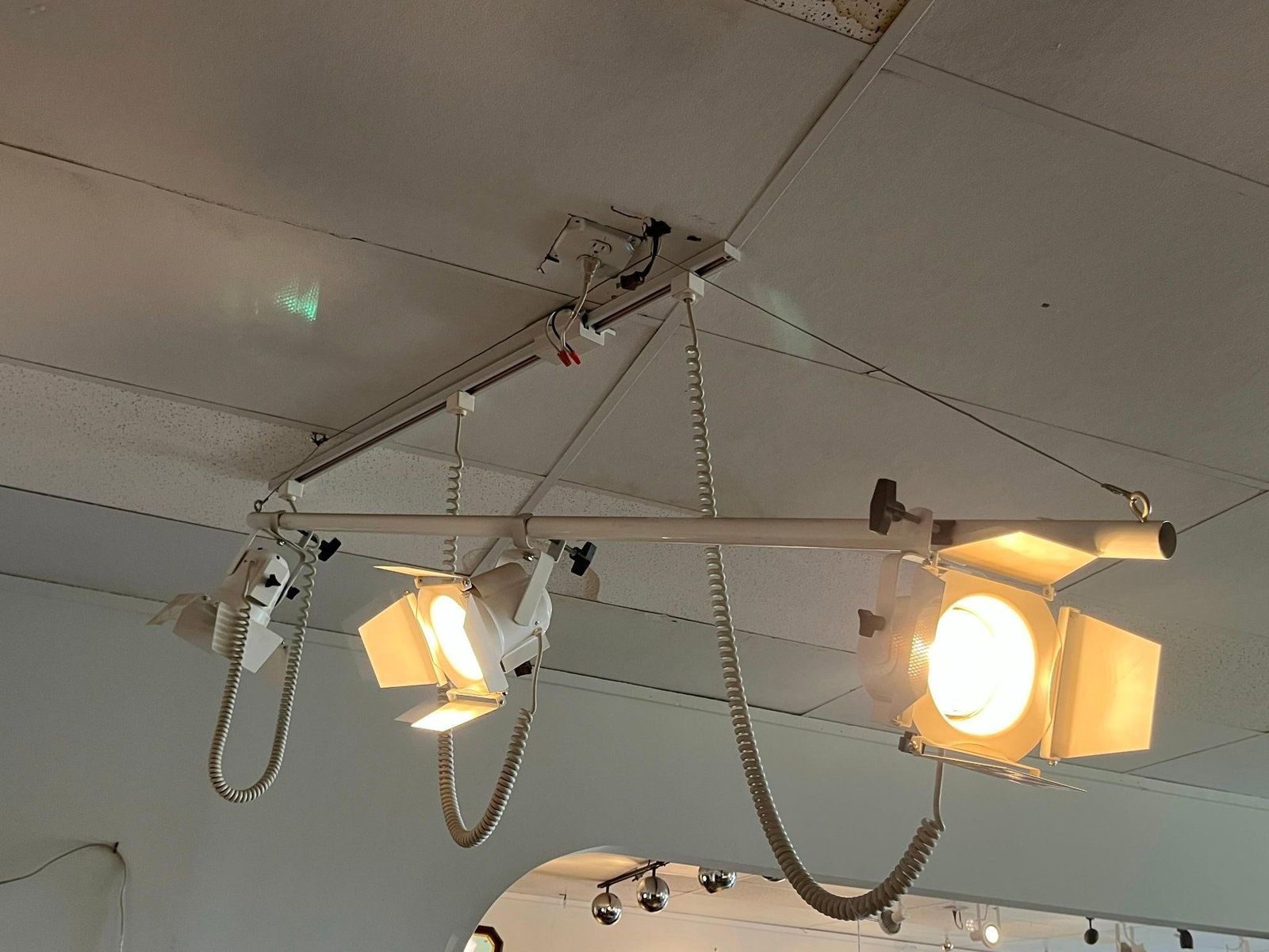 Triple Head Track Light w/ Barn Doors: A trio of industrial style studio track lighting heads mounted on a single horizontal suspension bar. Each of the three cylindrical lamp housings is constructed of metal with a semi-gloss white