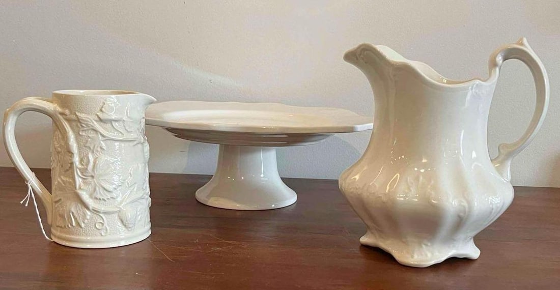 Spode & J.G. Meakin Ceramic Tablewares: This collection consists of three white glazed ceramic tablewares featuring two pitchers and a pedestal cake stand. The first item is an English earthenware pitcher featuring high-relief molded