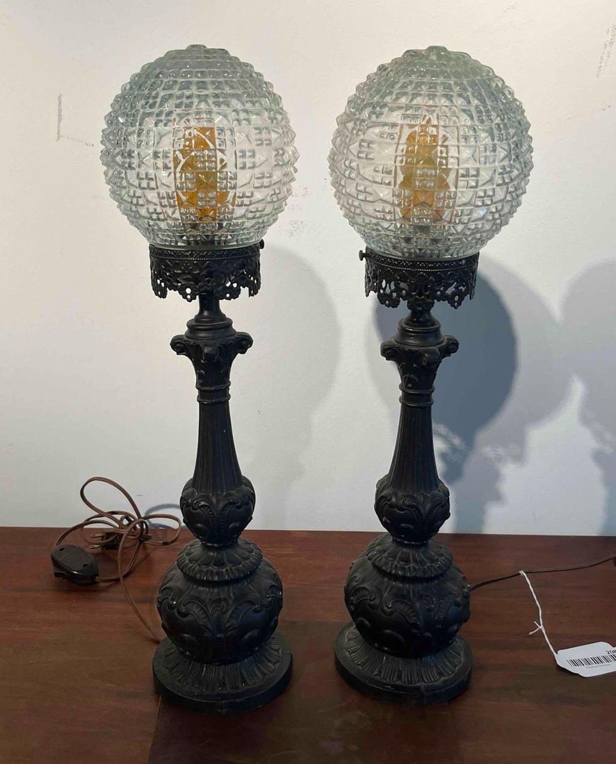 Pair Neo-Classical Metal Lamps w/ Glass: A pair of decorative table lamps featuring ornate cast metal bases with a matte black finish and clear pressed glass shades. Each lamp is designed in a Neo-Classical revival style with a fluted centra