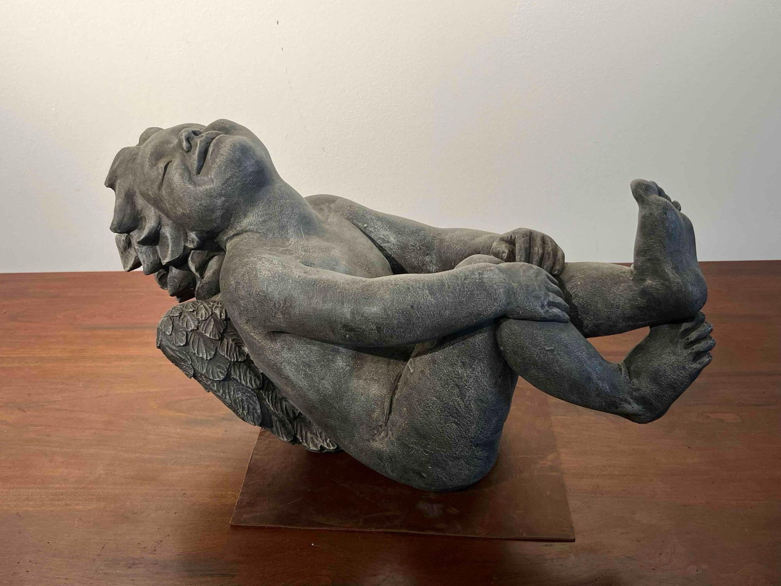 Cast Stone Reclining Cherub Sculpture: This large decorative garden sculpture depicts a winged cherub or putto in a playful, reclined pose. The figure is modeled lying on its back with its knees drawn toward its chest and its head tilted b