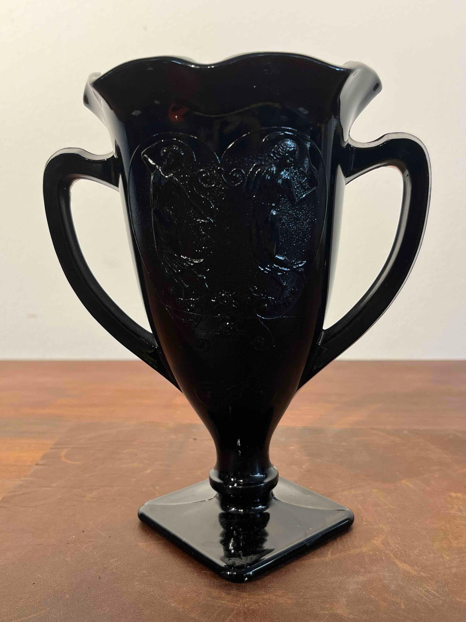 Black Glass Double-Handled Trophy Vase: This footed trophy-style vase is made from pressed black glass, which has an opaque appearance. The body is decorated with an embossed scene of two female figures in classical dress within a heart-sha