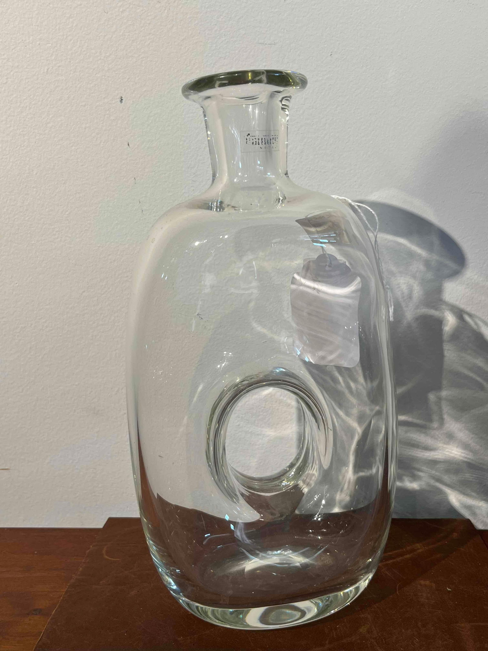 Polish Tarnowiec Pierced Donut Bottle: A Polish hand-blown clear glass bottle featuring a distinctive modern sculptural form. The vessel is constructed with a flattened ovoid body characterized by a large central circular piercing or torus