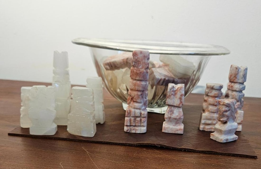 Mexican Onyx Chess Set in Glass Bowl: A set of chess pieces hand carved from two varieties of stone. One group is crafted from a translucent pale green and white stone, likely onyx or calcite, while the opposing group is made from a