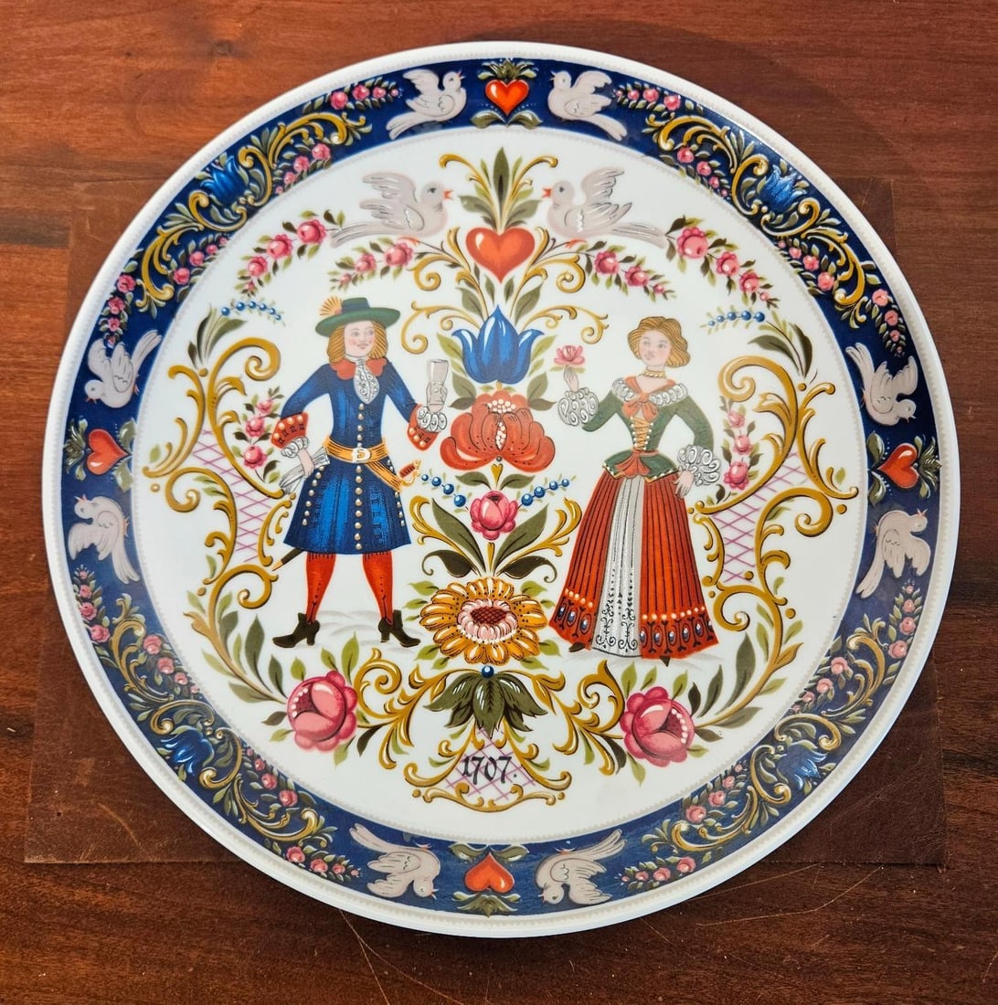 Kurt Hammer Porc. Folk Art Wall Plate: This circular decorative wall plate is constructed from glazed white porcelain. The piece features a multi-color transfer-printed scene in the style of traditional folk art. The central motif depicts
