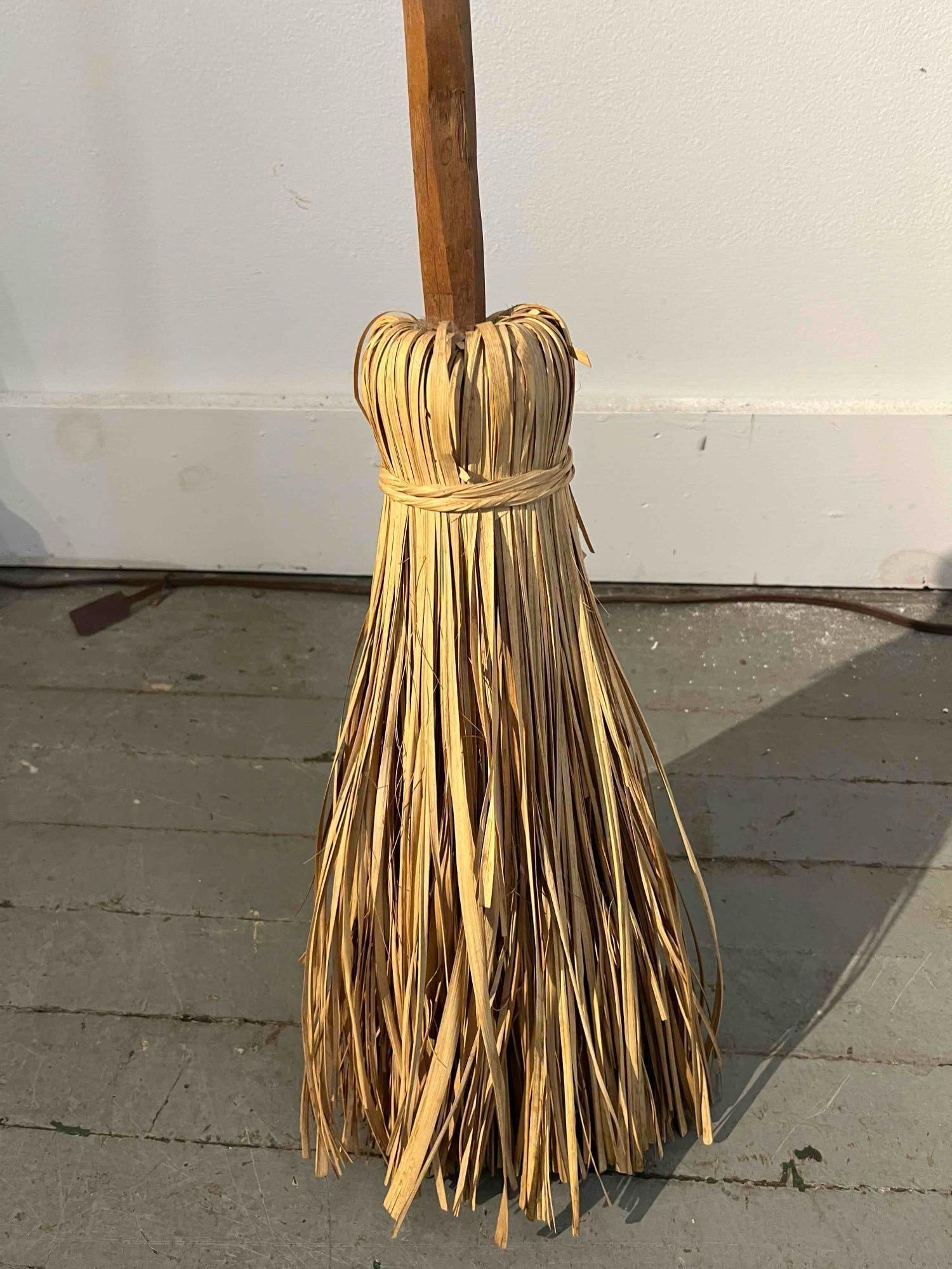 Besom Broom w/ Hand-Carved Wood Handle: A handmade artisanal besom broom featuring a hand-carved natural wood handle with a faceted, pointed top. The bristle section is constructed from flat-striated natural fibers, such as split palm or ra