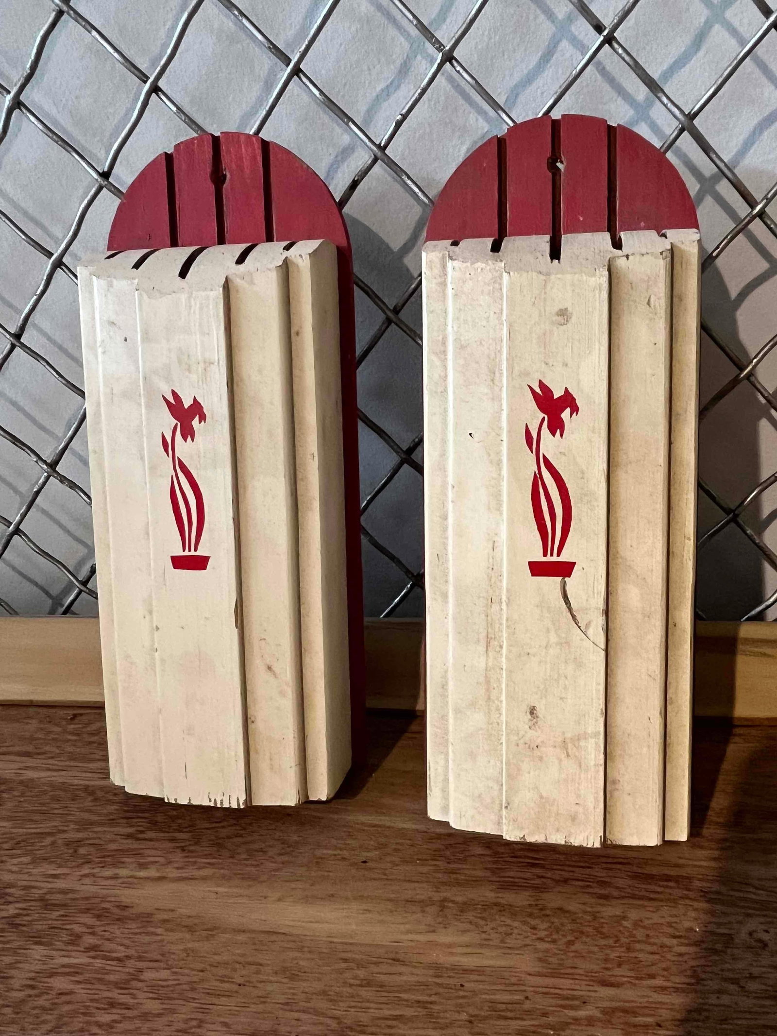 Pair Wall-Mount Wood Knife Holders: A pair of wall-mounted knife holders constructed from wood. Each unit features an arched backboard painted red with vertical slots at the top. The front blocks are painted in a cream color with