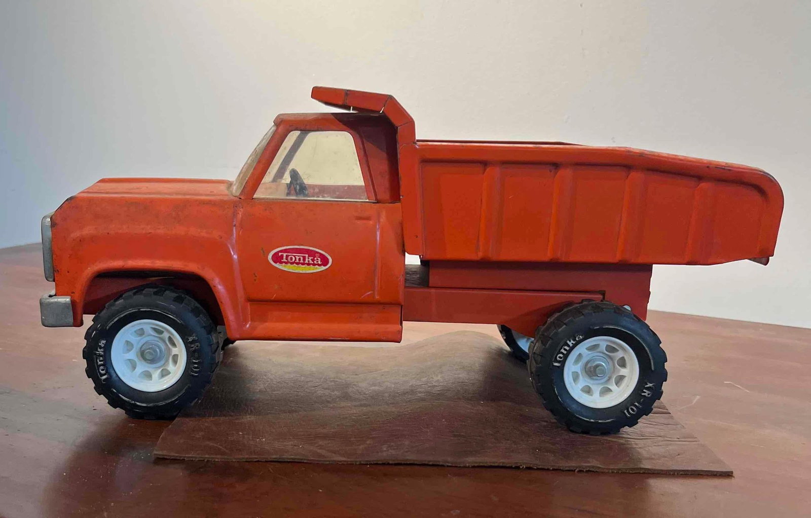 Tonka Pressed Steel Orange Dump Truck: A pressed steel dump truck manufactured by Tonka. The vehicle features a bright orange finish on its pressed steel cab and dump bed, accented by a silver-toned metal front bumper and grille assembly. 