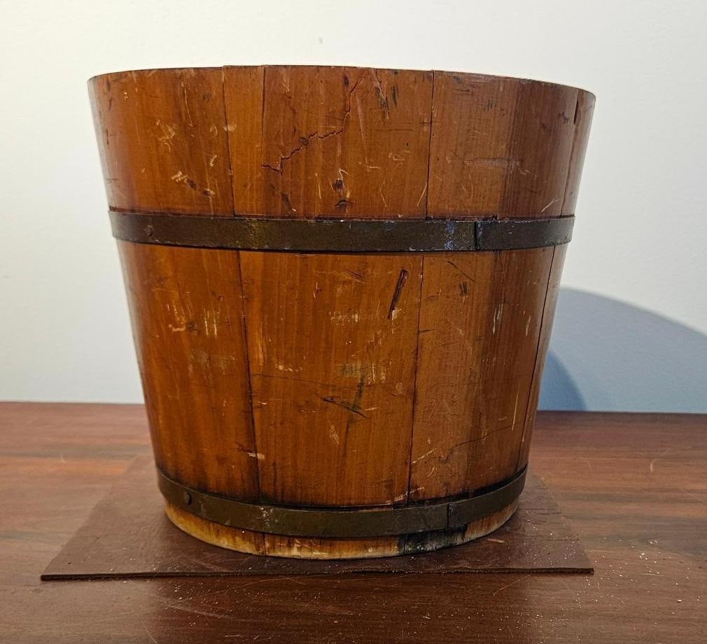 Primitive Wood Stave Sap Bucket: A wooden bucket of tapered cylindrical form, featuring traditional coopered construction. The vessel is composed of vertical wooden staves secured by two flat metal hoops. The wood exhibits an aged, d