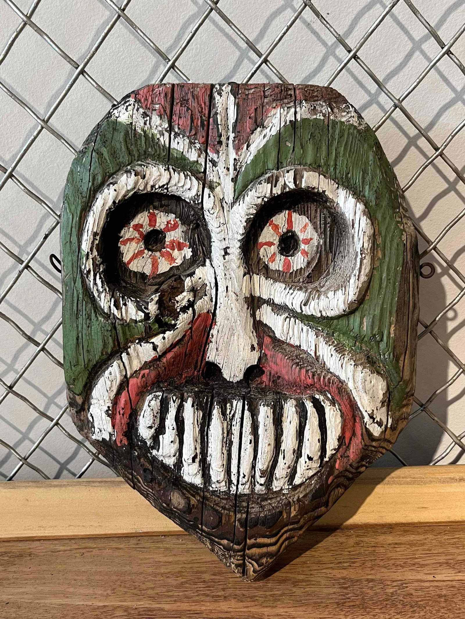 New Guinea Tiki Carved Wood Wall Mask: This hand-carved wood wall mask is crafted from a weathered, heart-shaped piece of salvaged lumber, exhibiting natural grain and vertical fissures. The face features large, deeply recessed circular ey
