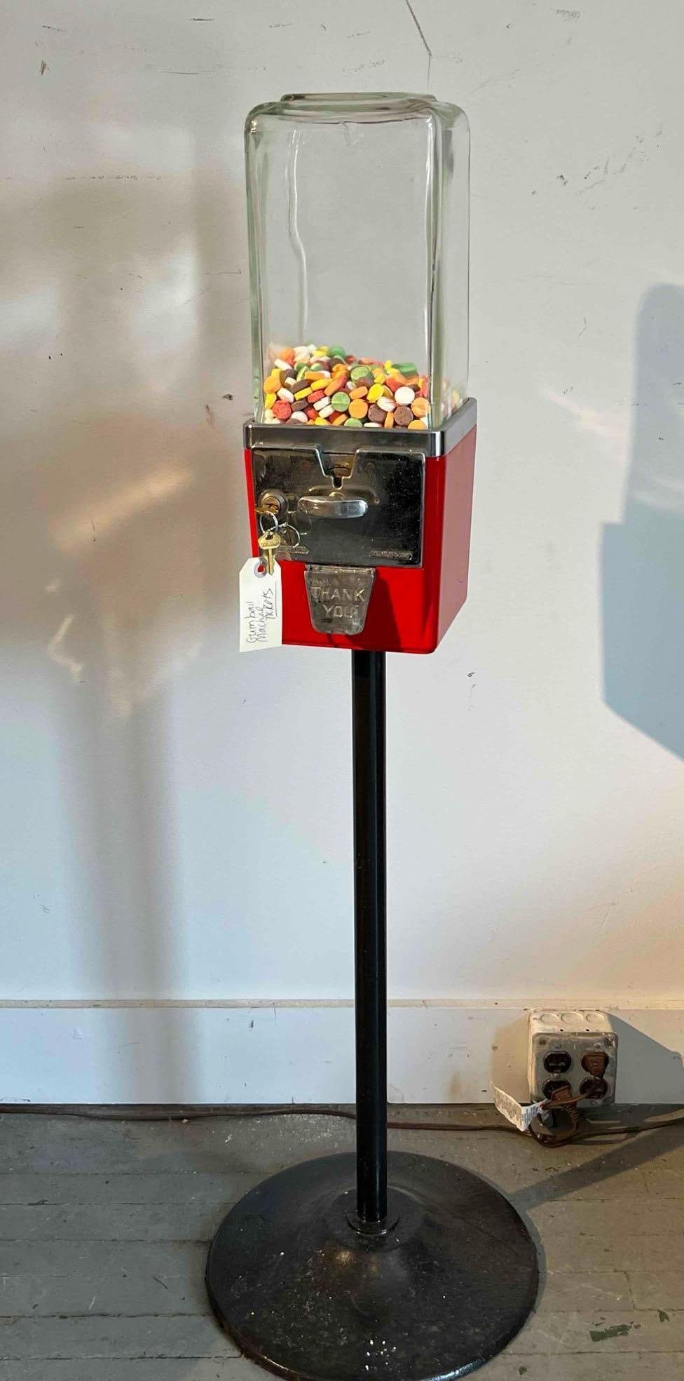 Pedestal Coin-Op Gumball Machine: This coin-operated vending machine features a floor-standing design with a rectangular red enameled metal body mounted on a black-painted tubular metal pedestal stand. The unit is topped with a heavy-