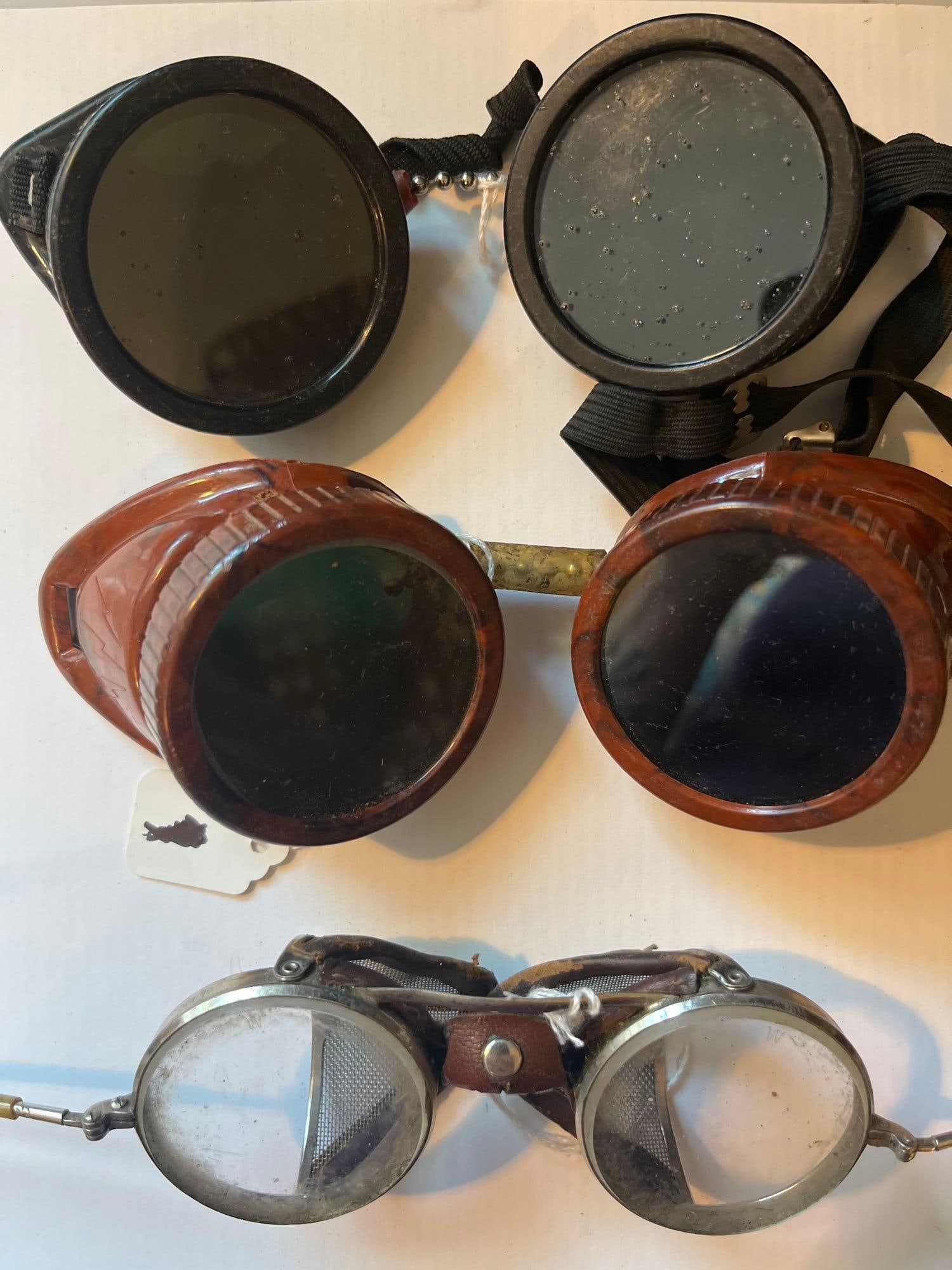 3 Pairs Safety & Welding Goggles: A trio of industrial safety goggles likely dating from the mid-20th century. The first pair consists of molded black hard rubber or plastic cup goggles designed for welding or metalwork. They feature 