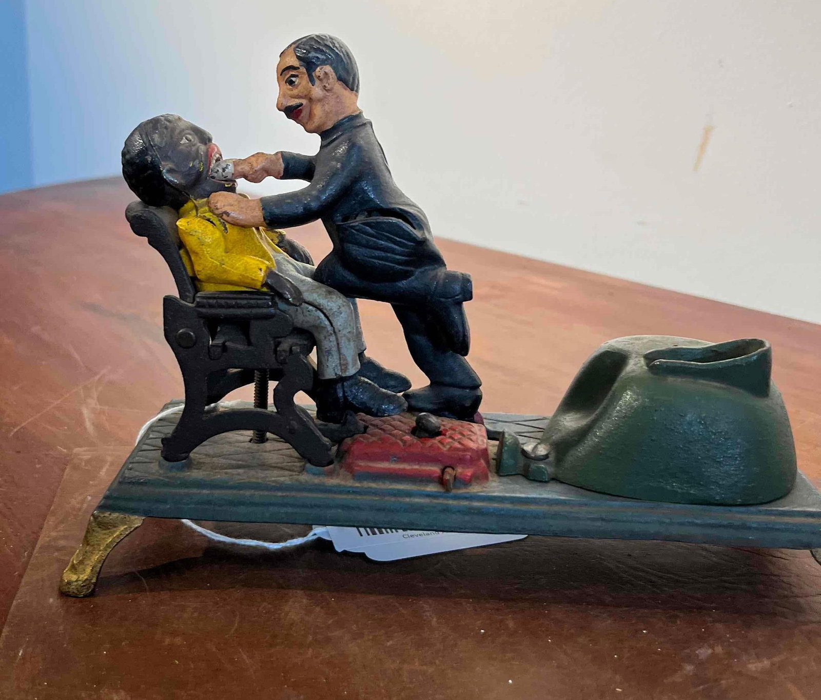 The Dentist Mechanical Bank: A cast iron mechanical bank featuring two polychrome figures. The scene depicts a dentist standing over a seated patient wearing a yellow shirt and gray trousers. The dentist wears a black suit and ho