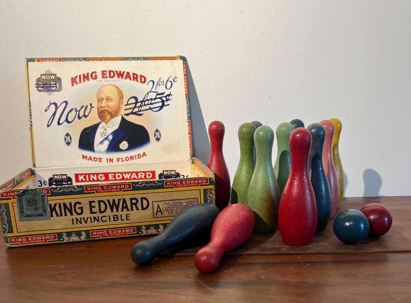 12 Wood Skittles & Balls, K. Edward Box: A set consisting of twelve turned wood pins and two balls, presented in a period cigar box. The pins and balls are finished in various colors, including red, green, blue, yellow, and pink. The pieces 