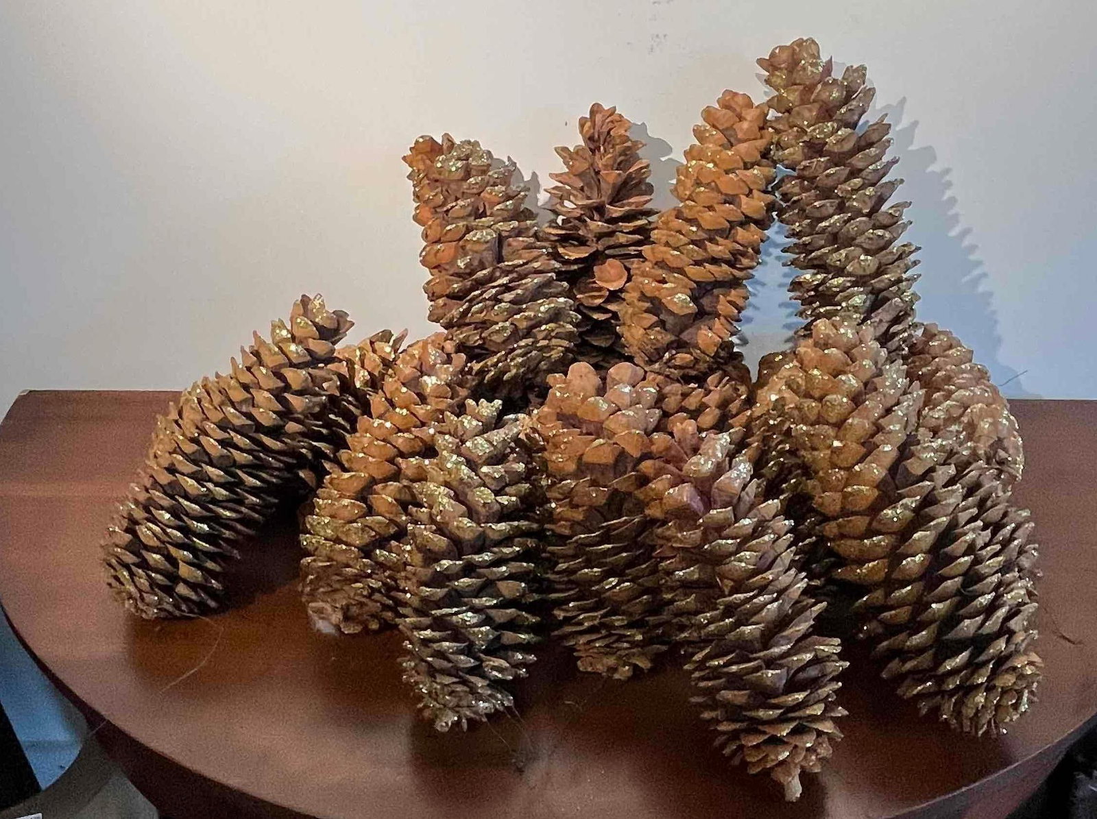 Gold-Tipped Pine Cone Collection: A collection of approximately 12 large, elongated natural pine cones. Each cone is composed of overlapping woody scales that have been embellished with gold-toned metallic glitter applied to the tips.