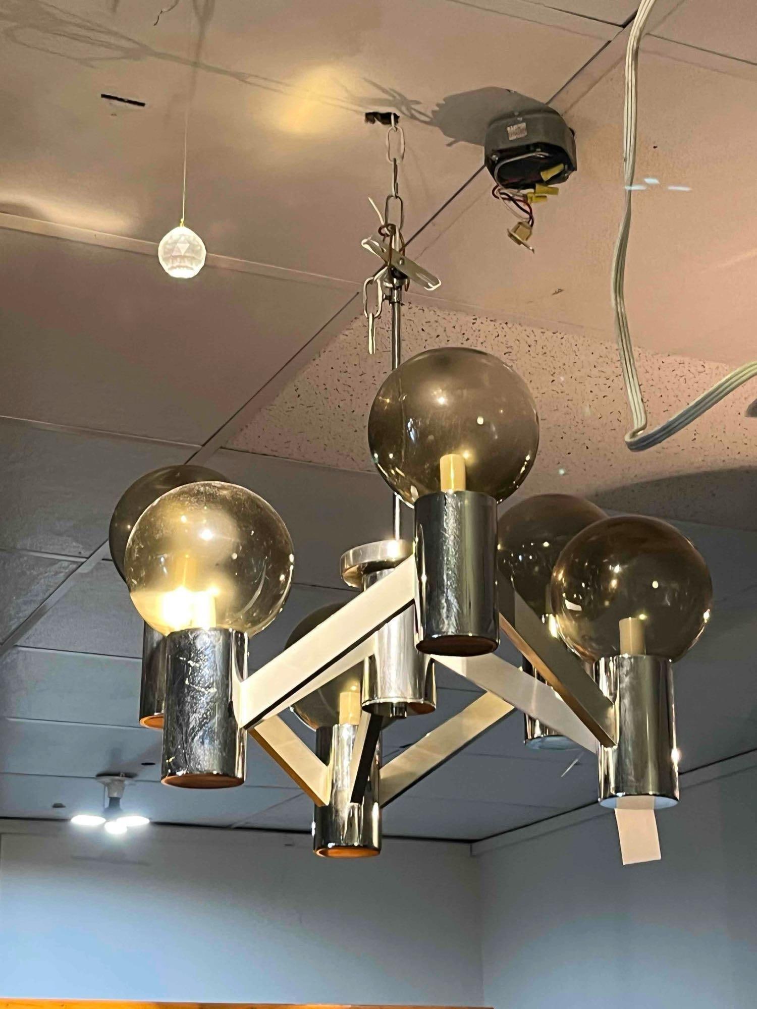 Mid-C. Chrome & Smoked Glass Chandelier: A six-light chandelier designed in the mid-century modern or Space Age aesthetic. The fixture features a chrome-plated metal frame consisting of a central vertical support rod and six rectangular hori