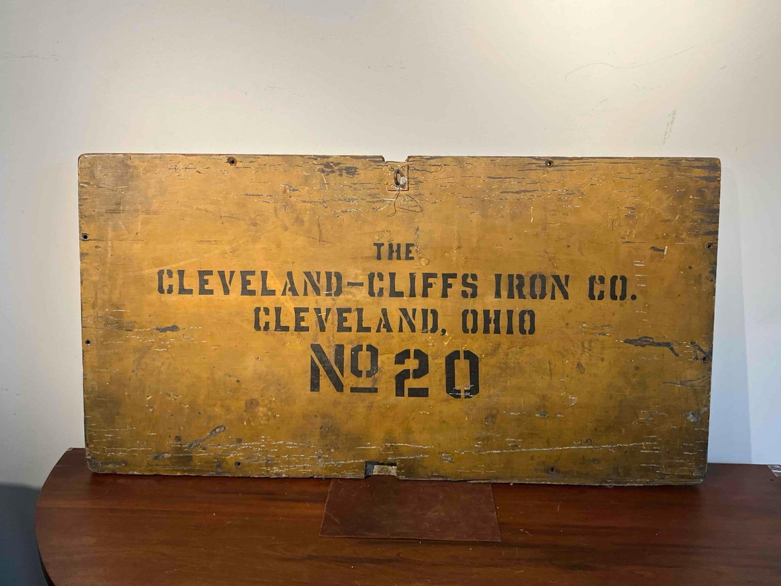 Cleveland-Cliffs Iron Co. Wooden Sign: An industrial stenciled wooden sign or crate panel for The Cleveland-Cliffs Iron Co. The rectangular board is finished in an ochre yellow paint with black stenciled lettering. The text is arranged in