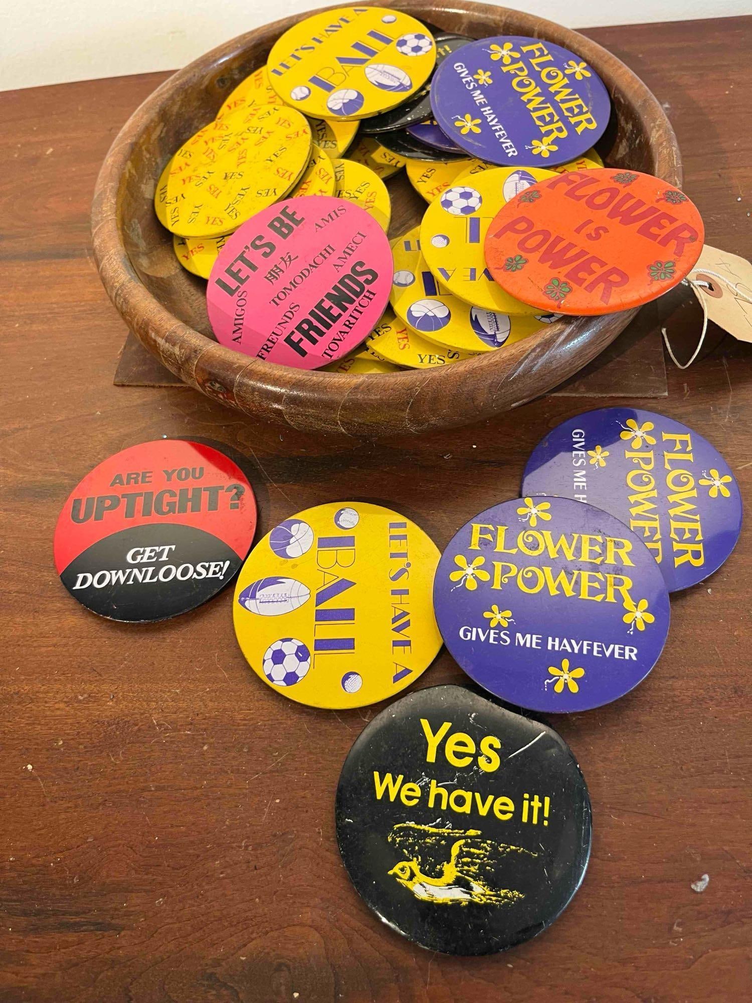 Novelty Pinback Buttons & Wood Bowl: An assortment of large-format novelty pinback buttons from the circa 1960s-1970s era, presented in a turned wooden bowl. The buttons are constructed of lithographed tin and feature slogans and
