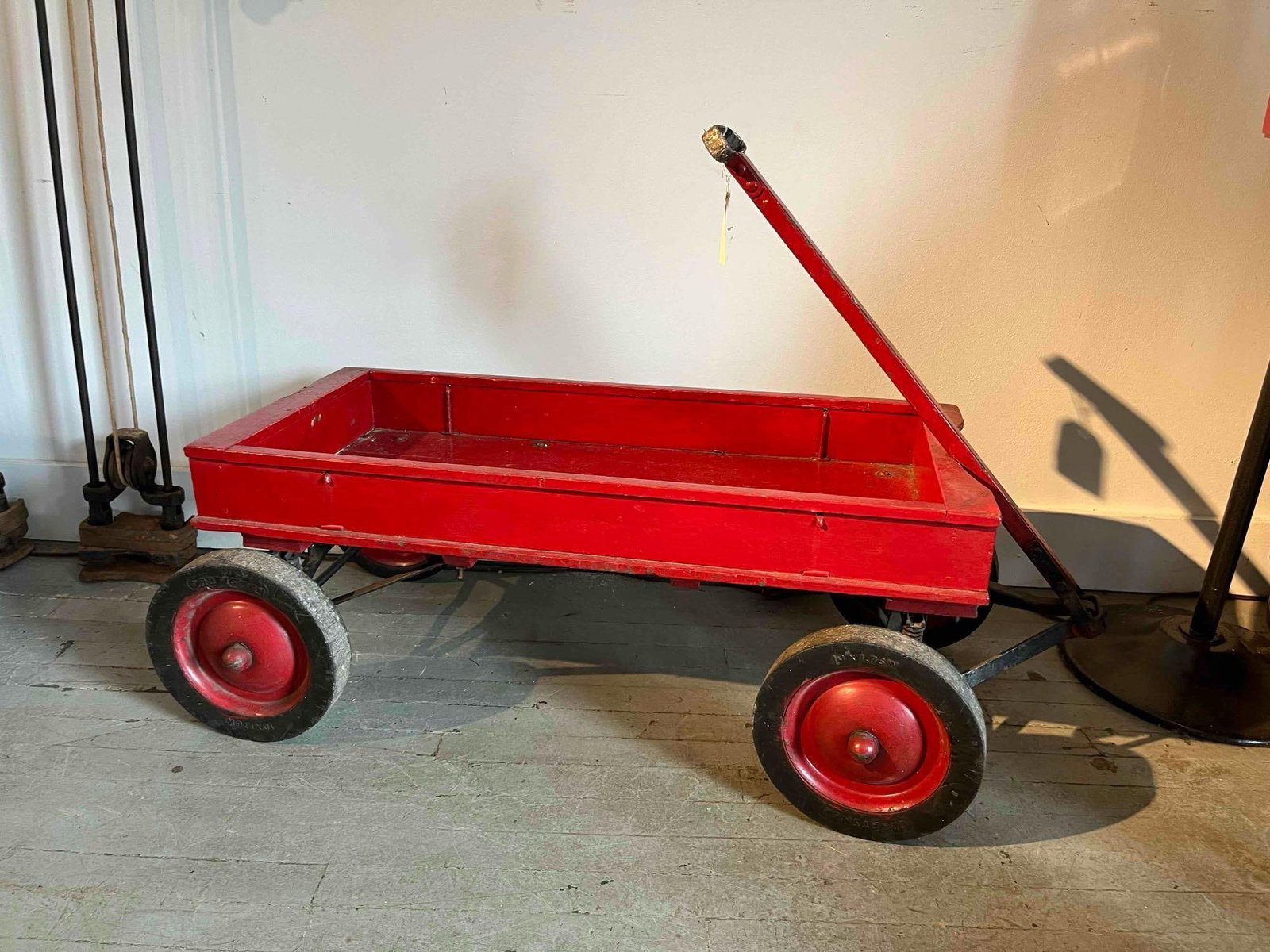 Roadmaster Wooden Wagon w/ Coil Spring: A child's pull wagon featuring a rectangular wooden bed and a straight wooden handle, both finished in red paint. The wagon is mounted on a black-painted metal chassis with a steerable front axle. A n