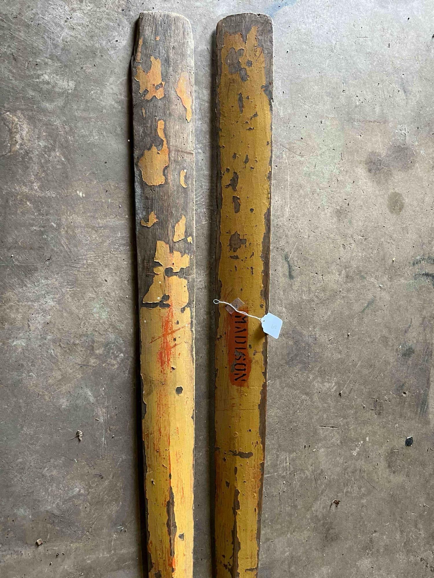 Pair of Local Madison Yellow Wood Boat Oars: A pair of long wooden boat oars constructed from carved wood with a tapered shaft and narrow blades. The oars feature a distressed yellow painted finish throughout. Each blade displays a rectangular