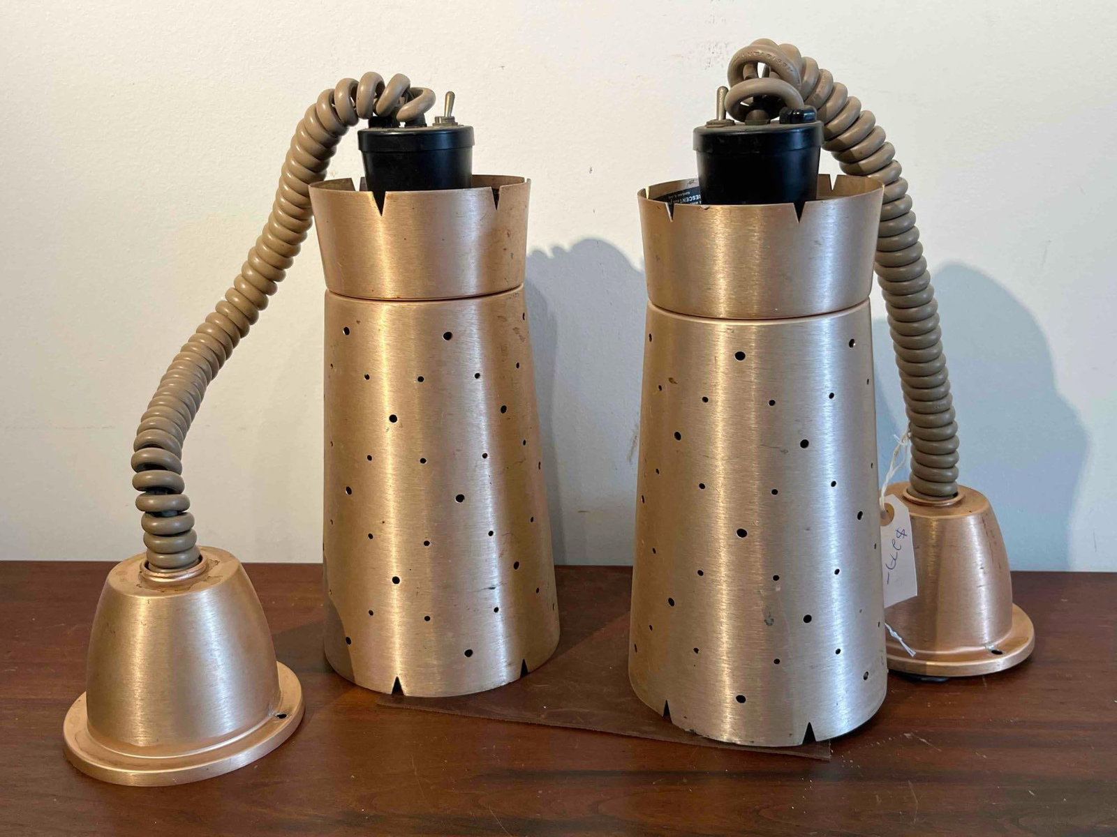Pair Retractable Copper Heat Lamps: A pair of matching industrial retractable heat lamps featuring conical spun aluminum shades with a copper-tone finish. The shades are detailed with a repeating pattern of circular perforations