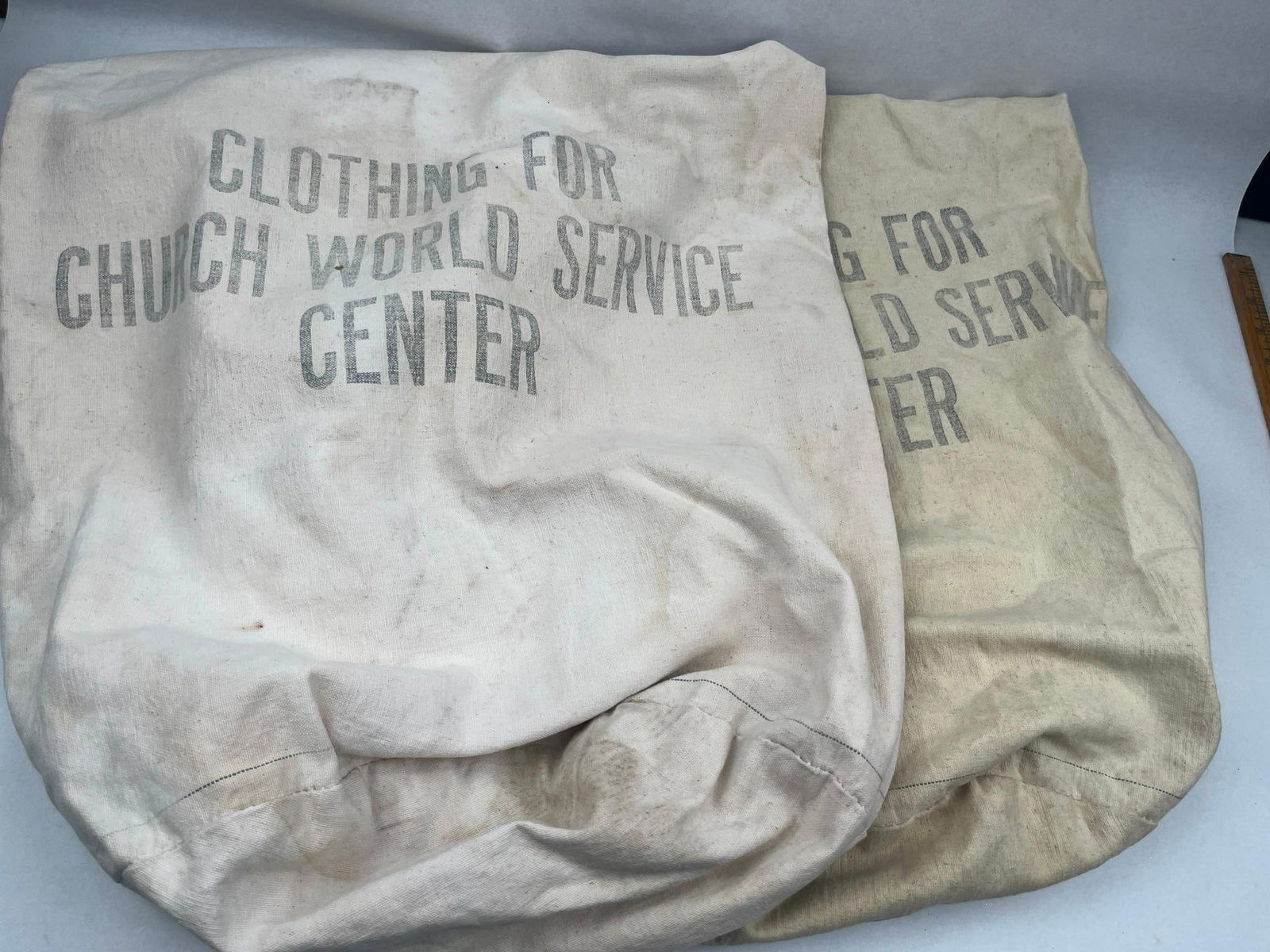 Pair Church World Service Donation Bags: A pair of heavy-duty canvas laundry-style donation bags used by the Church World Service. The bags are constructed from off-white and tan canvas duck cloth. Each bag features a drawstring closure at