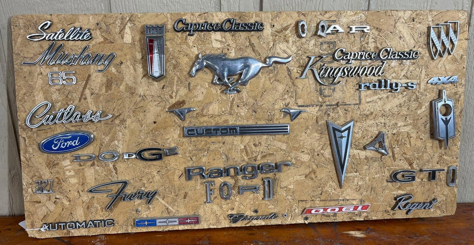 Vtg Ford Dodge & Buick Badges & Emblems: An assortment of approximately 28 vintage automotive emblems, nameplates, and mascots mounted on a rectangular oriented strand board. The collection predominantly features chrome-plated die-cast zinc
