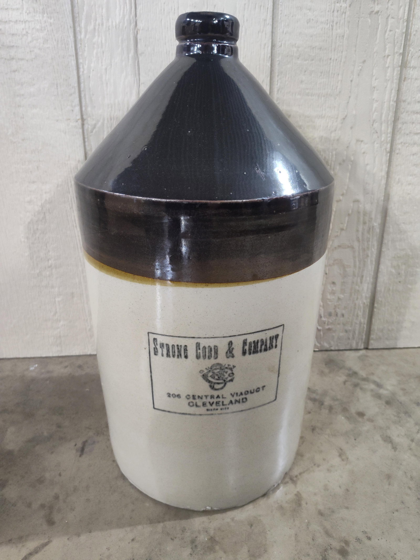 Strong Cobb & Co Two Tone Stoneware Jug: A two-tone stoneware shoulder jug featuring a strap handle. The vessel is finished with a dark brown slip glaze covering the neck, shoulder, and handle, over a cream-colored Bristol-glazed