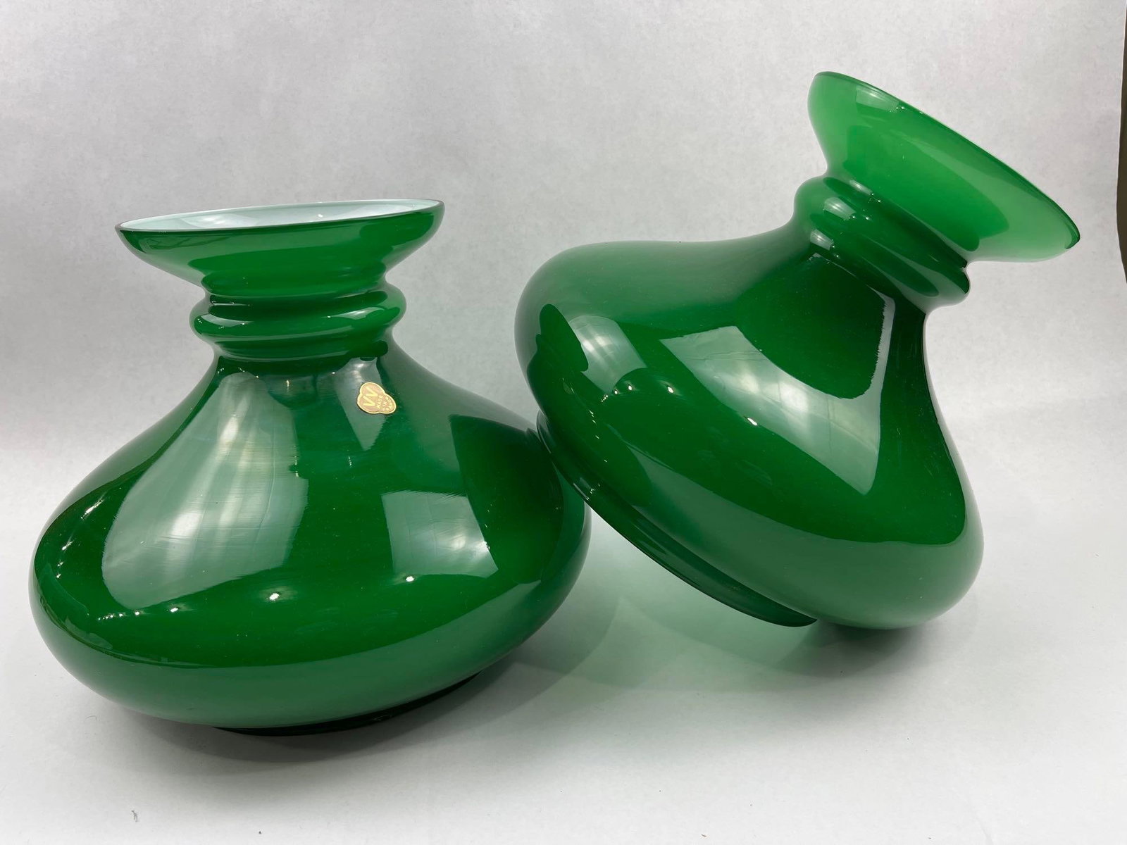 Pair French Cased Green Glass Shades: A pair of French cased glass lamp shades for use with student lamps, featuring a vibrant emerald green exterior and an opaque white interior. One shade bears a trilobed gold-toned foil adhesive label