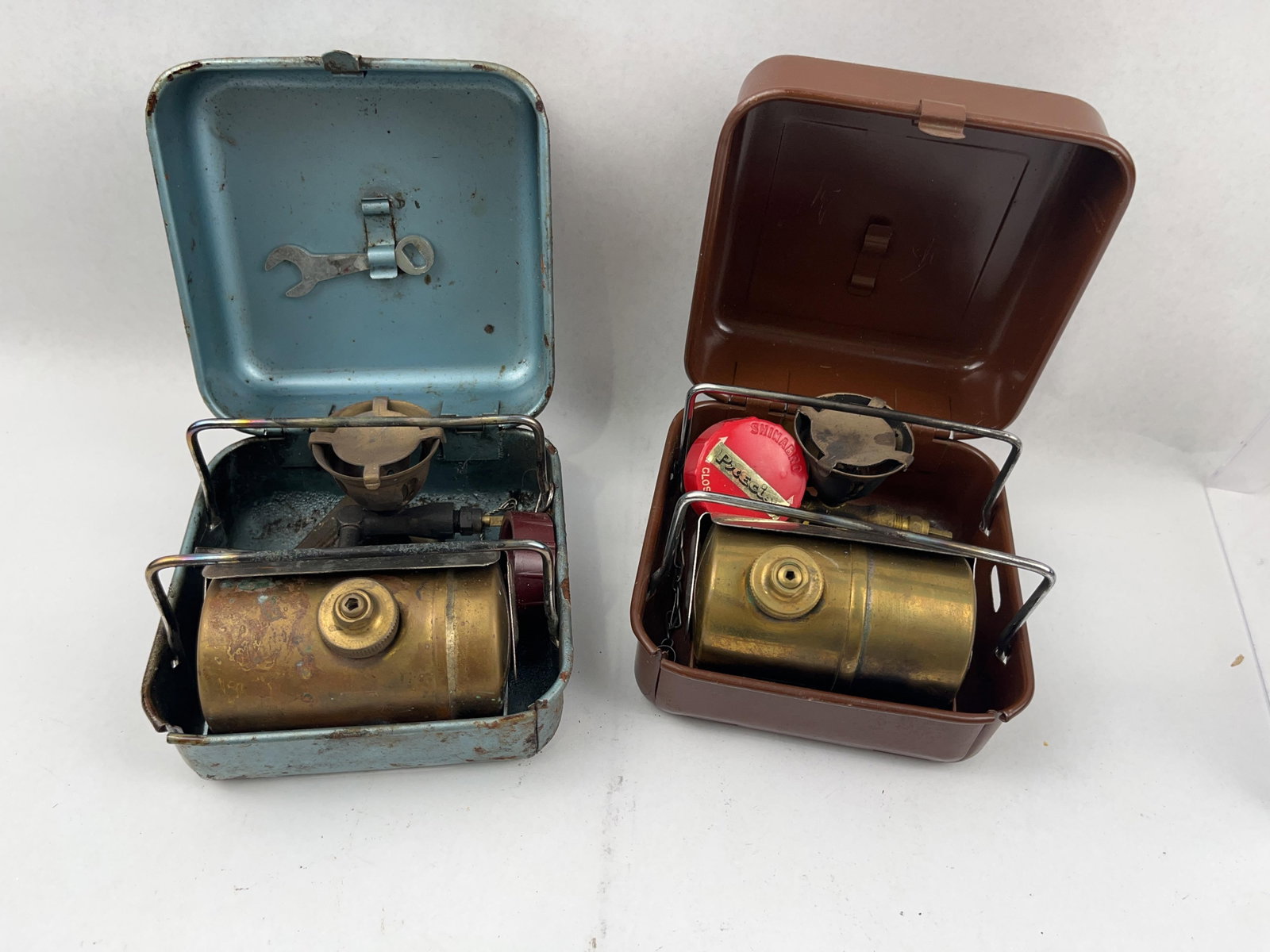 Pair Primus 8R & Precise Phoenix Stoves: This lot features two vintage portable gasoline camping stoves, each housed in its original metal carrying case. It is accompanied by a lid-mounted metal wrench and a control key with a maroon