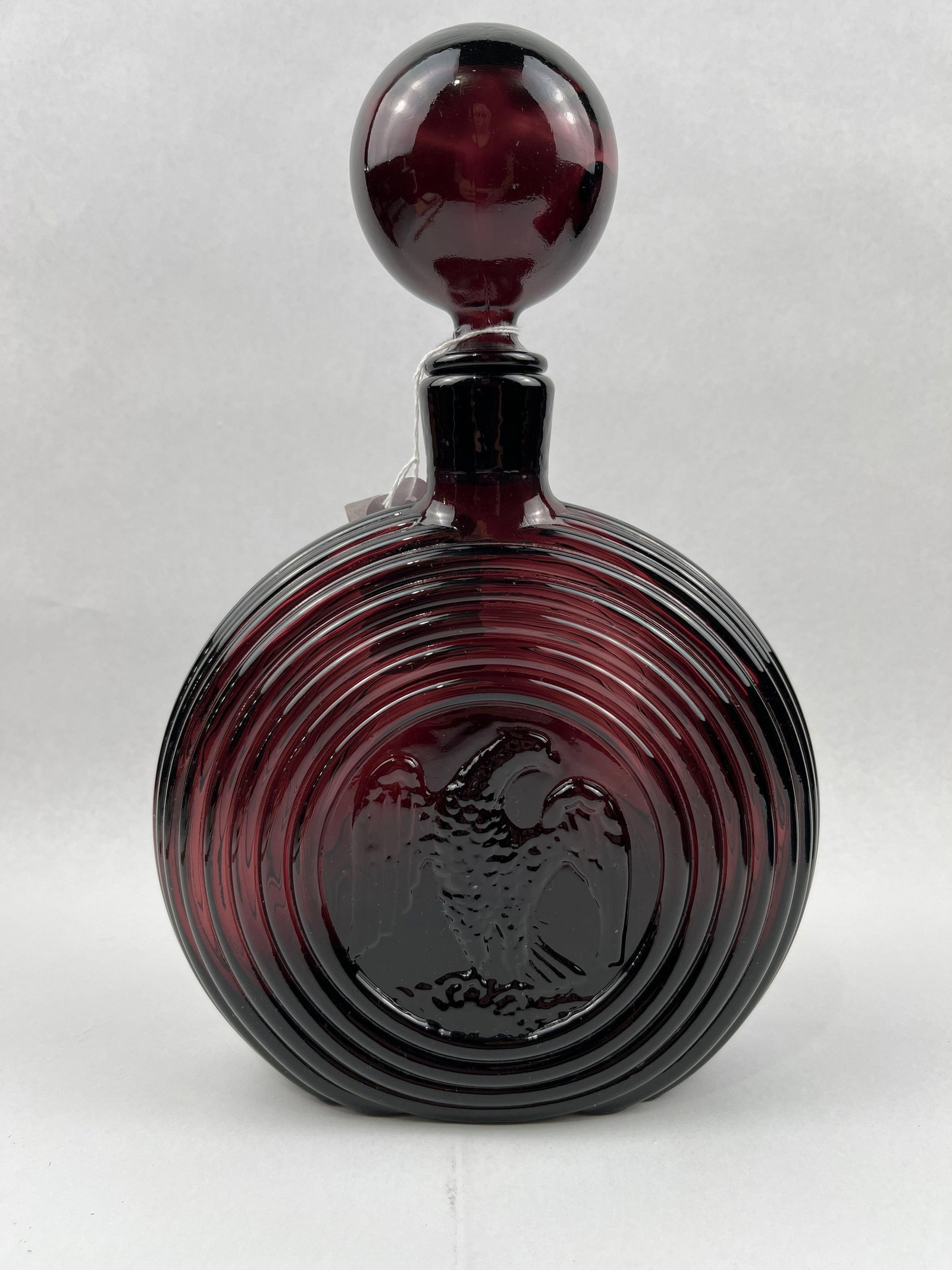 Ruby Red Glass Decanter w/ Eagle Motif: A mold-blown decanter in deep ruby red glass featuring a flattened circular form. The body is decorated with relief-molded concentric rings that frame a central circular medallion on each side. There