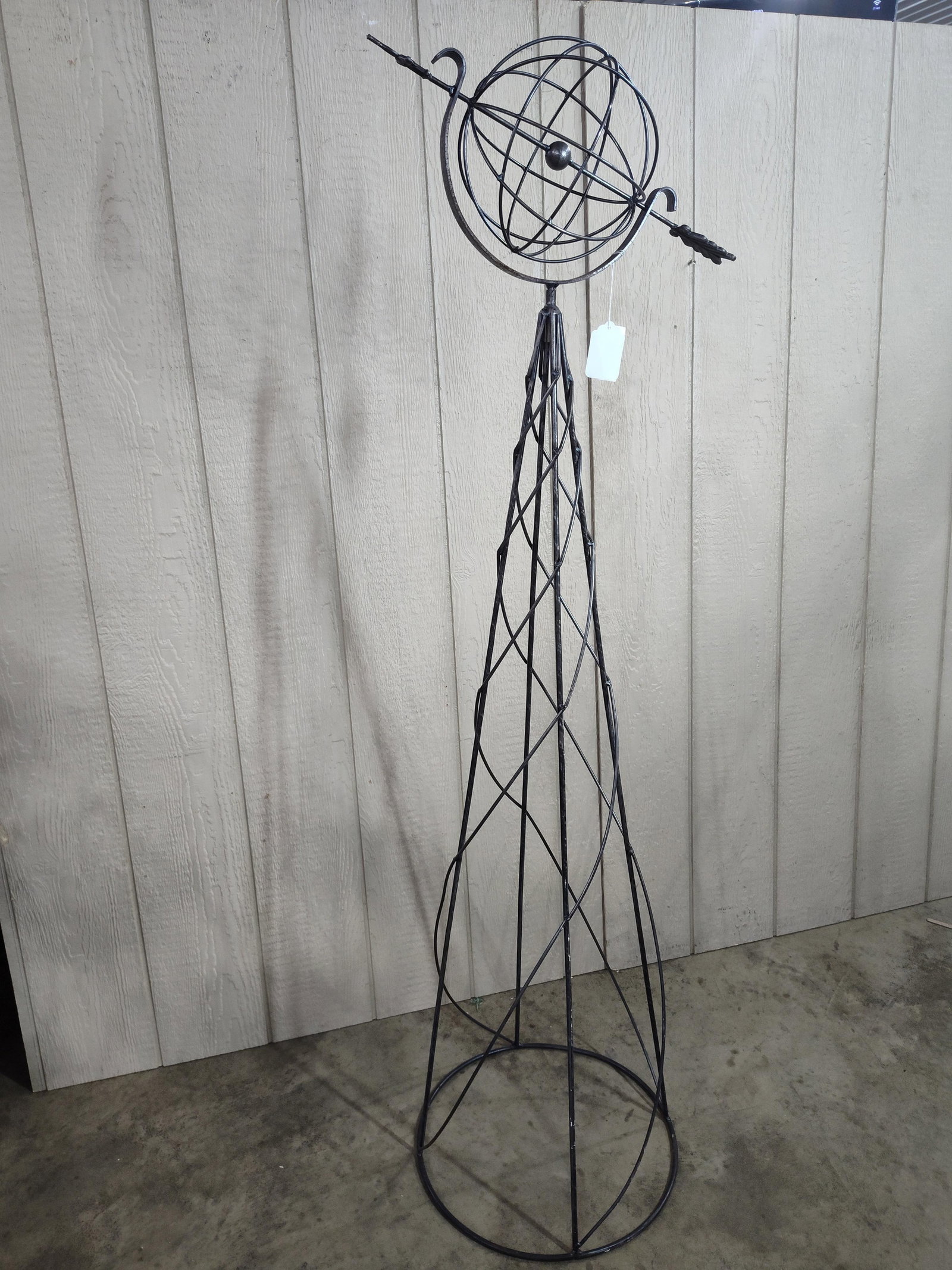 Wrought Metal Armillary Sphere Obelisk: A tall garden obelisk constructed of wrought metal with a dark weathered finish. The structure features a tapered openwork frame composed of spiraling and intersecting rods that form a geometric latti