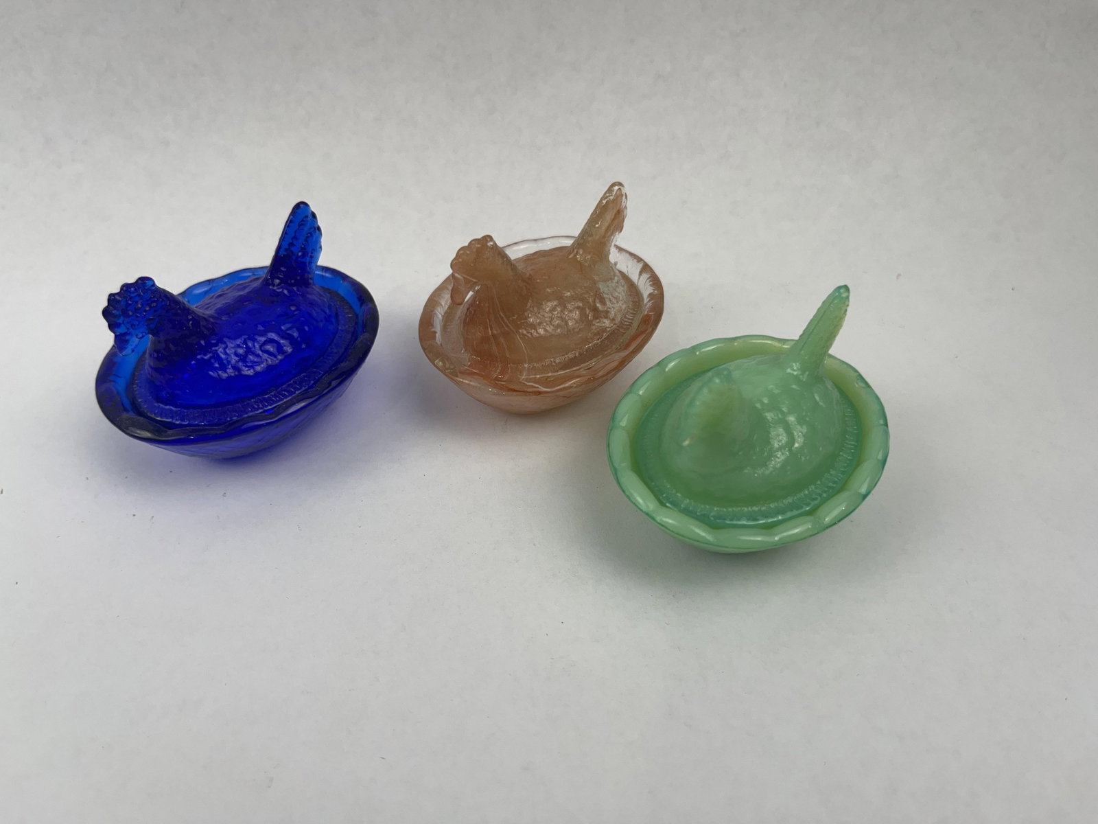 Trio Pressed Glass Hen on Nest Dishes: A collection of three miniature two-piece pressed glass covered dishes, each modeled in the classic hen on nest form. The group includes one dish in transparent cobalt blue glass, one in marbleized