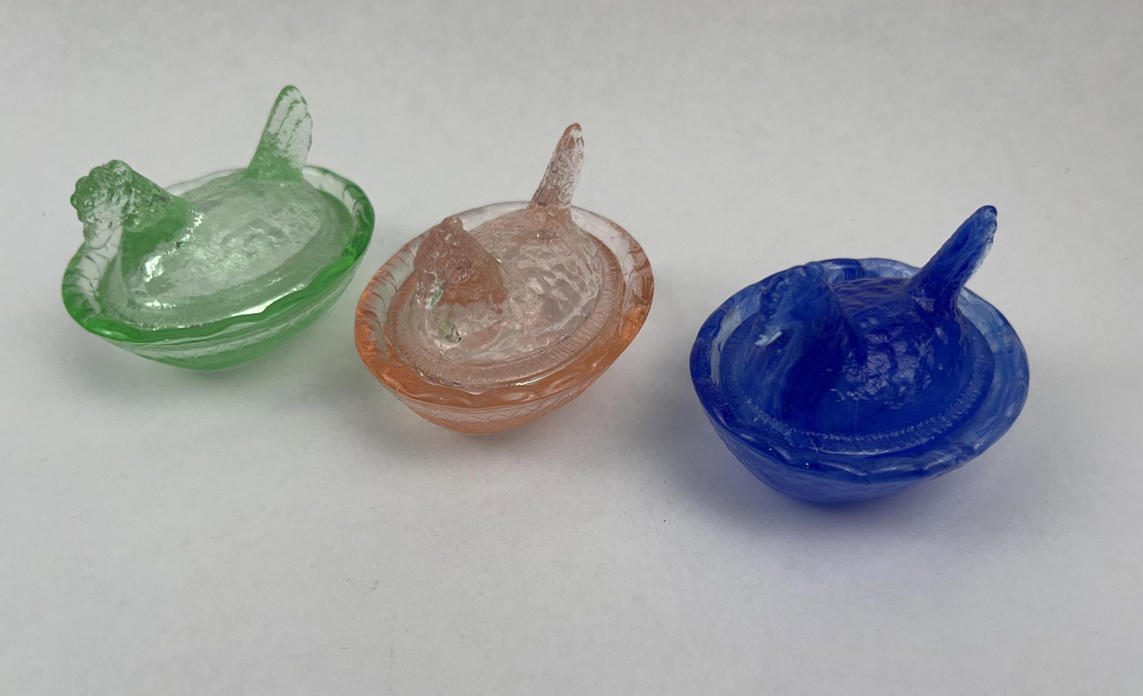 Trio Glass Hen on Nest Lidded Dishes: A trio of miniature pressed glass lidded dishes in the form of a nesting hen. The group features three distinct color variations: one in transparent lime green, one in transparent peach-pink, and one