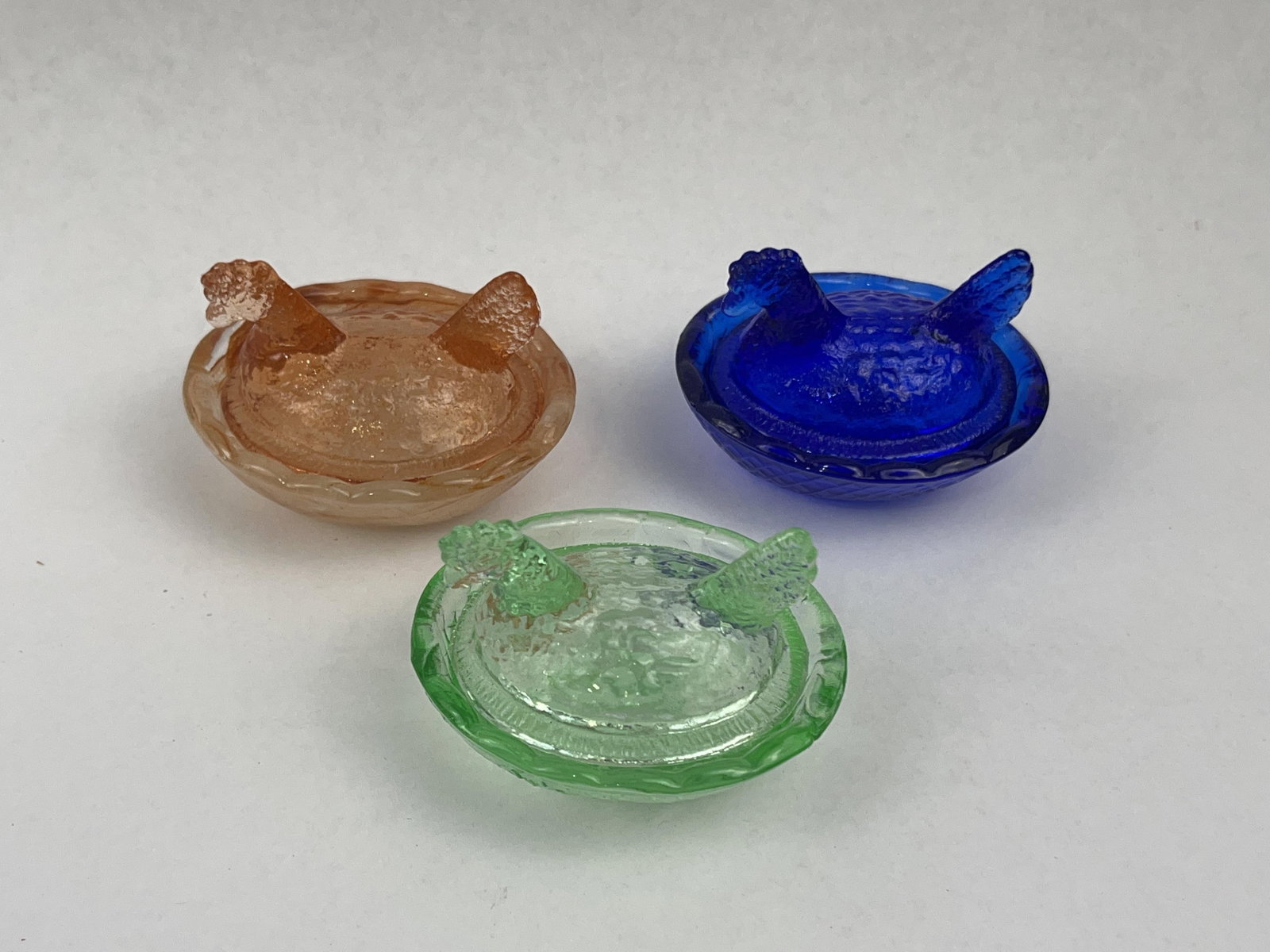 Trio Pressed Glass Hen on Nest Dishes: A group of three miniature pressed glass covered dishes, designed in the form of a hen on a nest, often utilized as individual salt cellars. Each piece comprises two parts: a lid modeled as a nesting