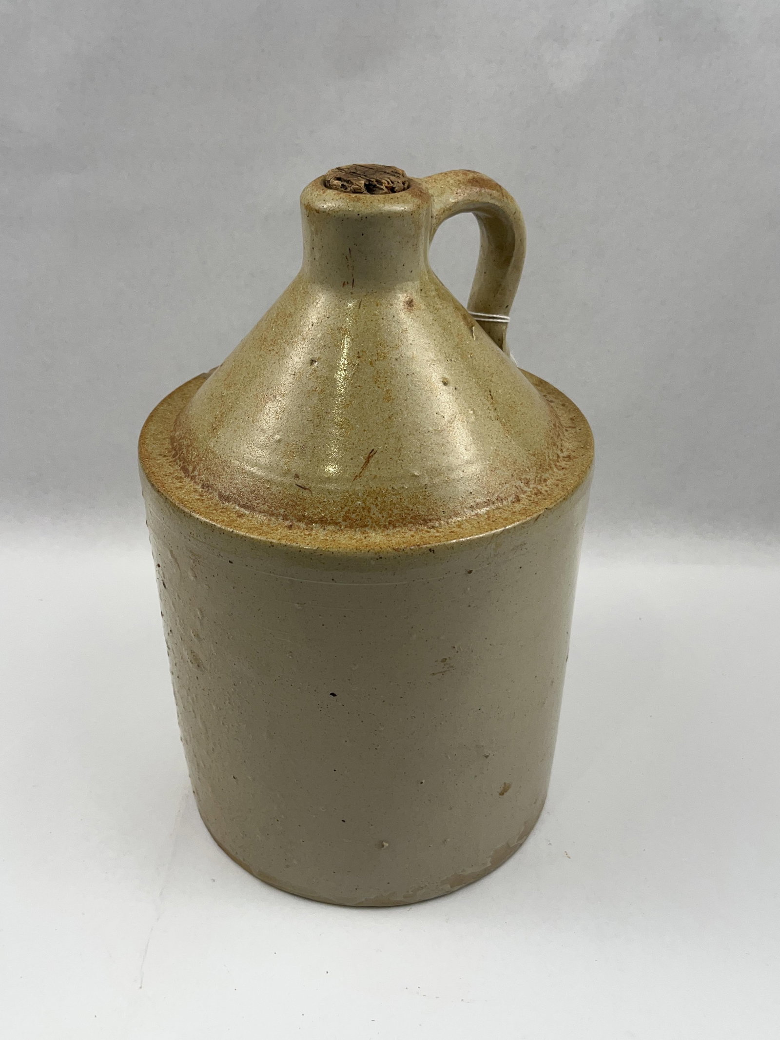 American Salt Glazed Stoneware Jug: A cylindrical salt glazed stoneware shoulder jug of utilitarian form. The vessel features a tapered shoulder, a short neck, and a single applied strap handle. The jug is undecorated and lacks cobalt