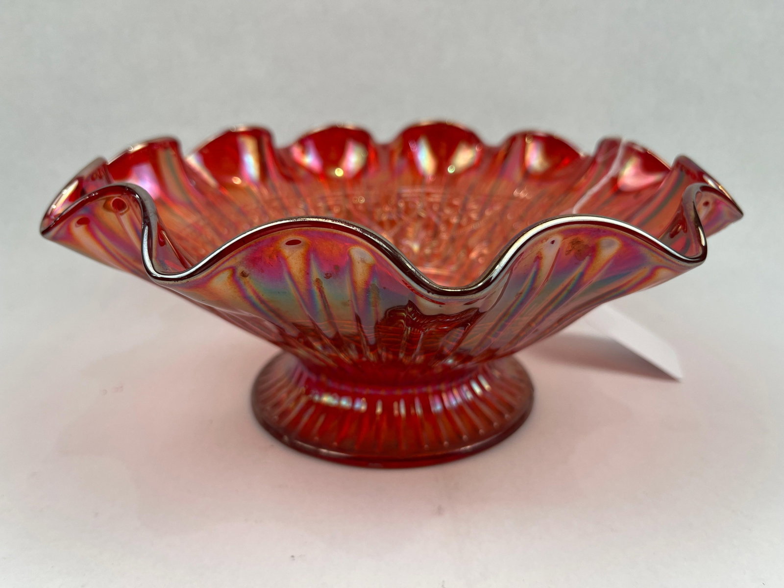 Fenton Red Carnival Glass Ruffled Bowl: A red carnival glass bowl featuring a deep iridescent finish with golden and purple highlights. The piece is constructed with a circular pedestal base showing vertical ribbing, supporting a wide,