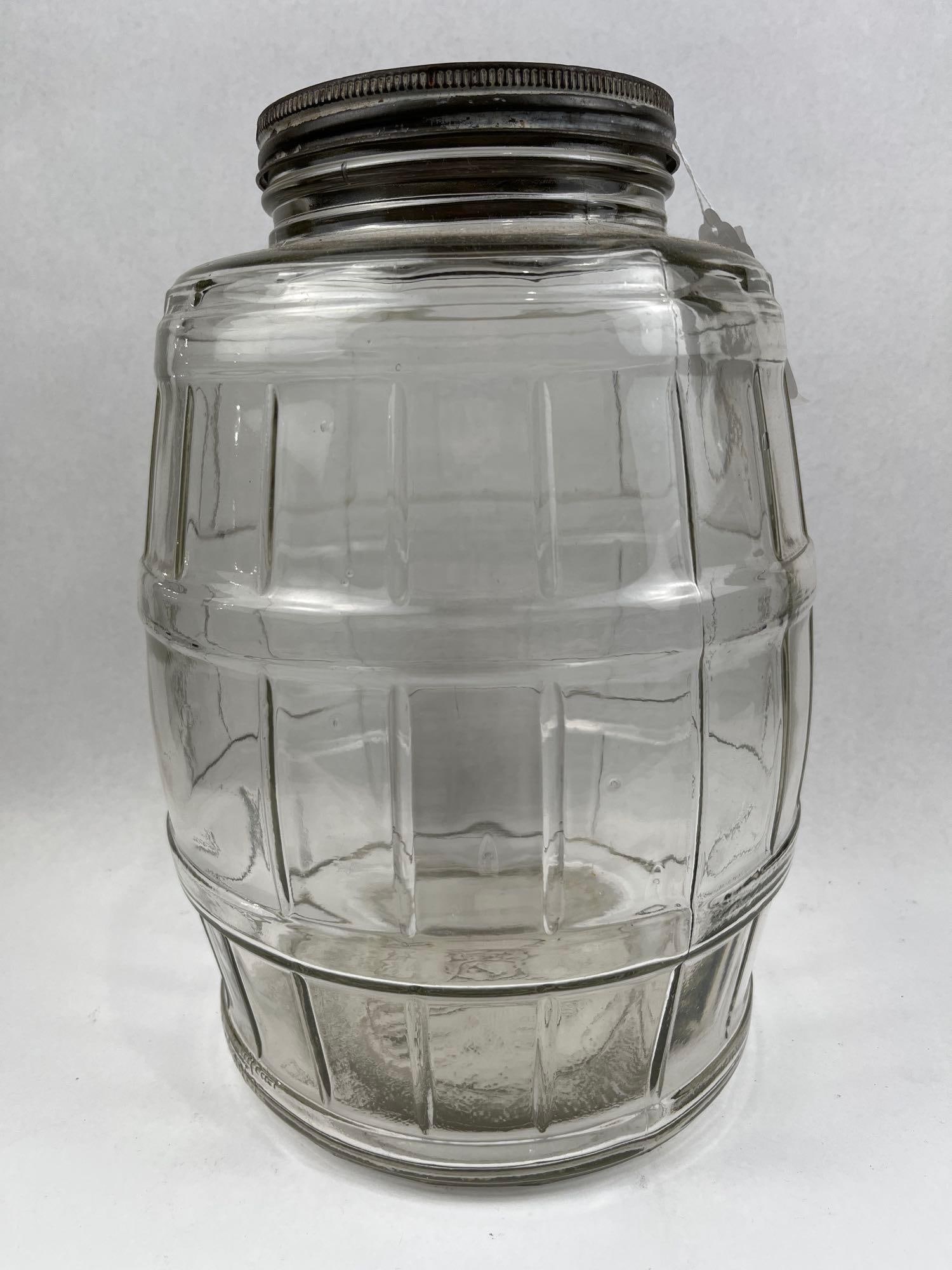 Molded Glass Barrel Store Counter Jar: A large molded clear glass storage jar designed in a barrel motif with vertical staves and horizontal reinforcement bands. The vessel is topped with a metal screw-on lid featuring a knurled rim. The