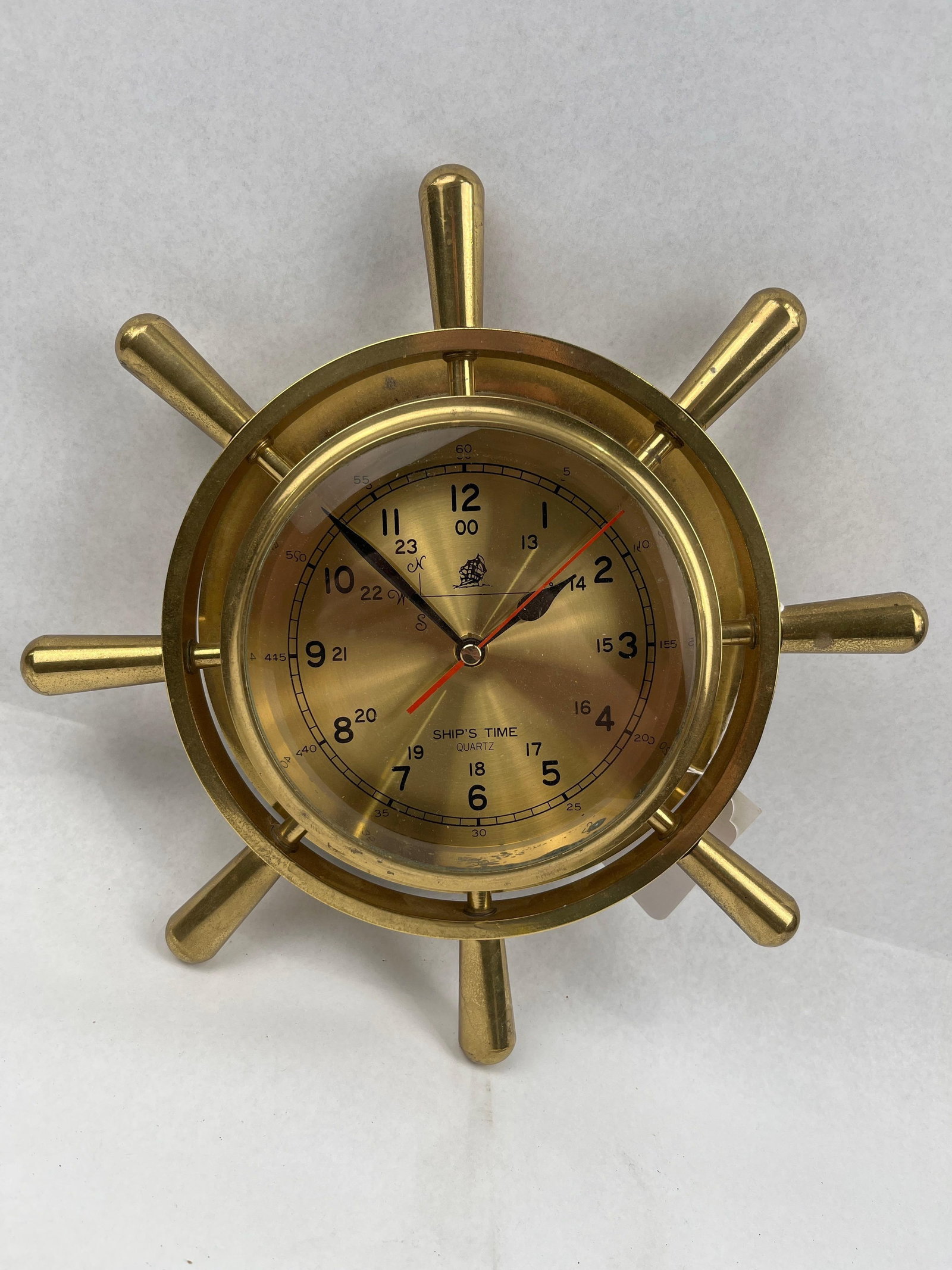 Brass Ship's Wheel Nautical Wall Clock: A brass-cased wall clock designed in the form of an eight-spoke ship's helm. The circular clock is set within a matching brass frame with eight radiating spokes. The dial is brass-toned and features