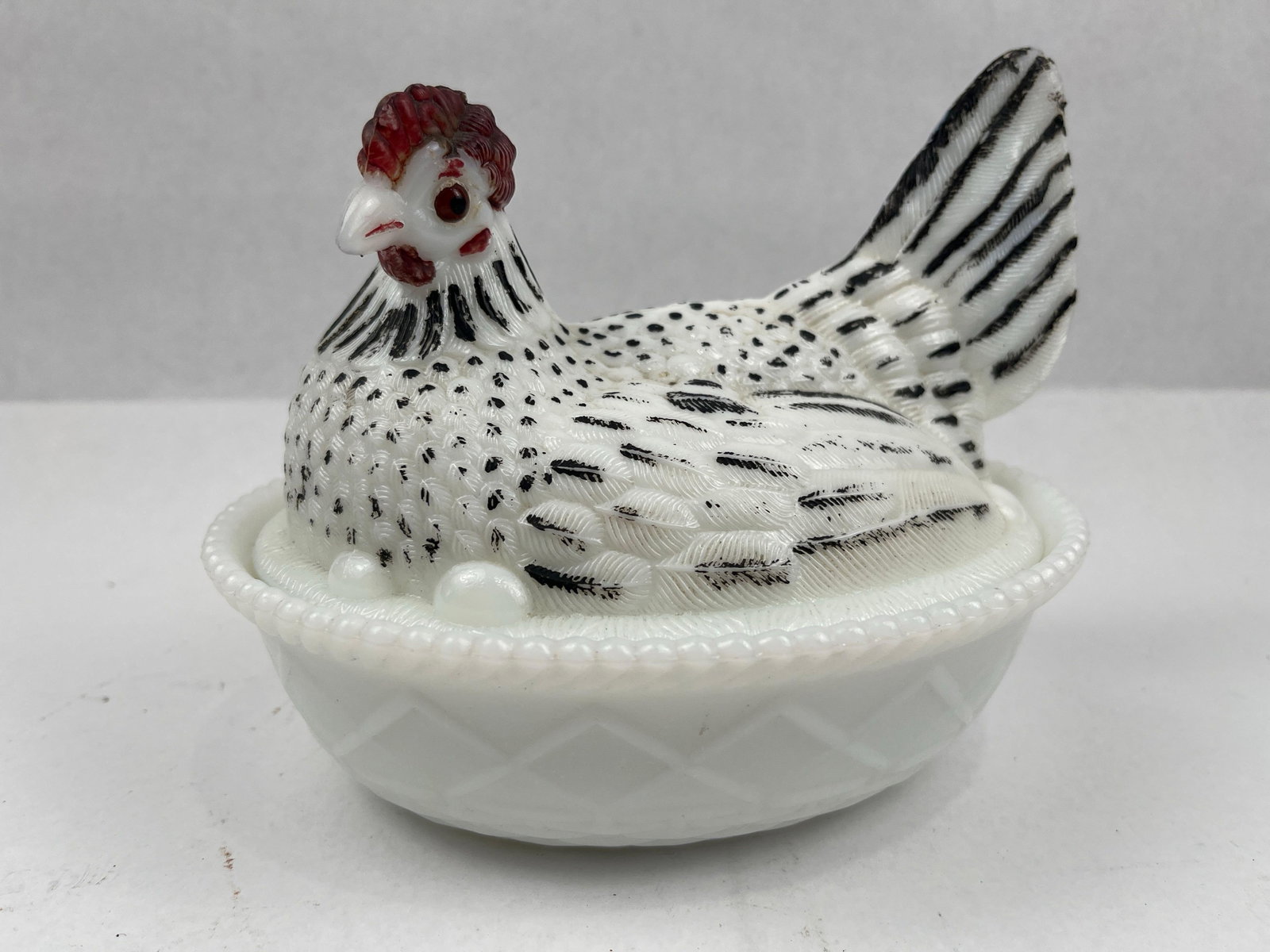Painted Milk Glass Hen on Nest Dish: A two-piece covered dish composed of opaque white milk glass. The lid is sculpted in the form of a nesting hen surrounded by three eggs at the base of the body. The hen features hand-painted black