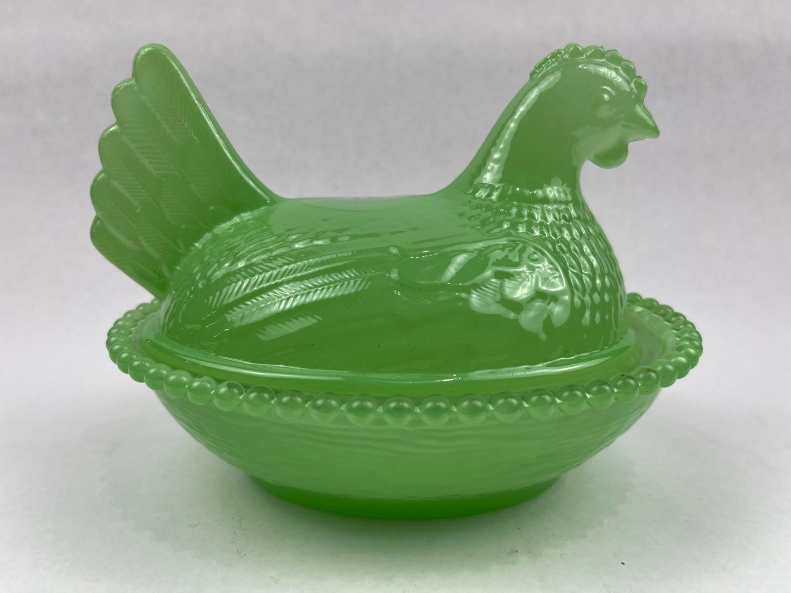 Opaque Green Pressed Glass Hen on Nest: This two-piece pressed glass covered dish is composed of a lid in the form of a nesting hen and a matching oval base, both rendered in a vibrant opaque lime green glass. The lid features detailed mold