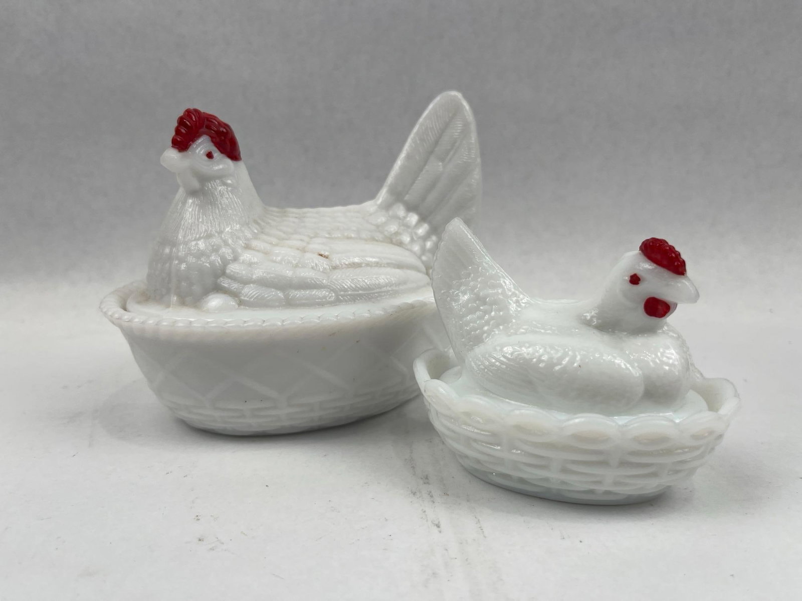 Pair Milk Glass Hen-on-Nest Dishes: Two opaque white milk glass covered dishes designed as hens seated on nests. The larger vessel features a hen with a textured feather pattern and a split tail, seated upon an oval base with a