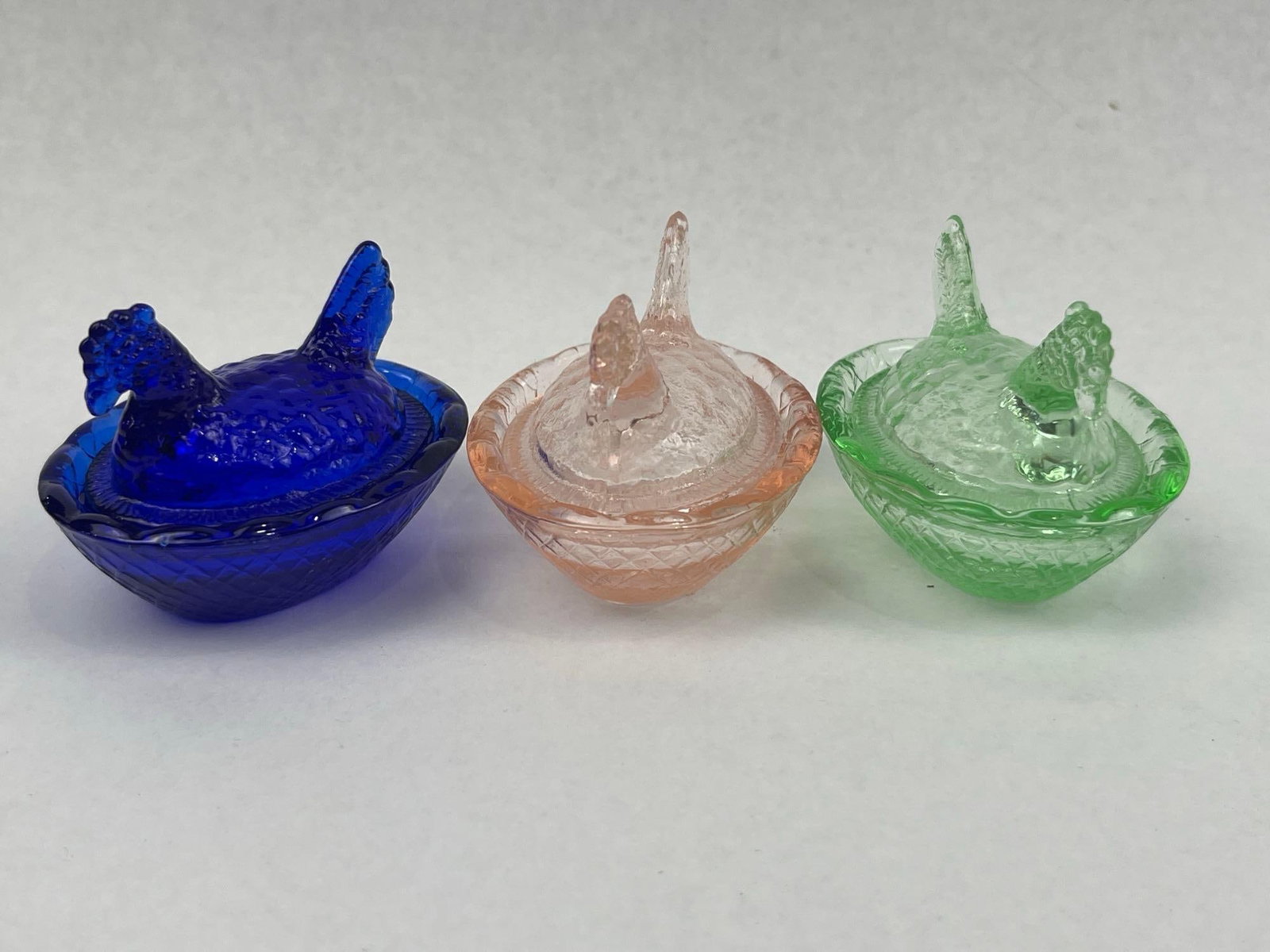 Trio Pressed Glass Hen on Nest Dishes: A trio of miniature pressed glass covered dishes in the form of hens on nests. The set comprises three separate colors: cobalt blue, light pink, and pale green. Each dish consists of a removable lid