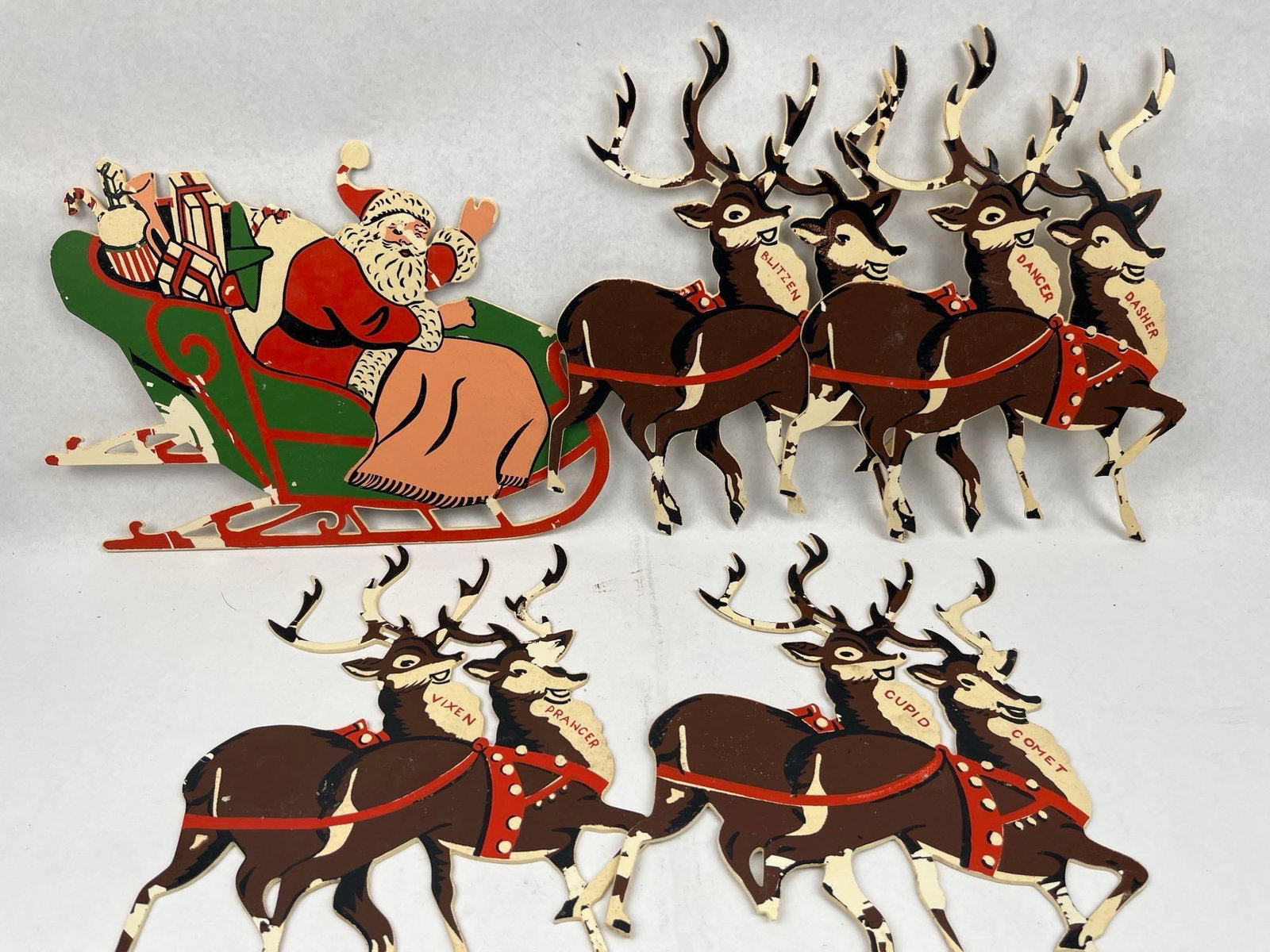 Hardboard Santa Sleigh & Reindeer: A group of vintage mid-century seasonal decorations featuring Santa Claus in a sleigh and several reindeer figures. These flat-profile cutouts are constructed from painted hardboard. The sleigh is