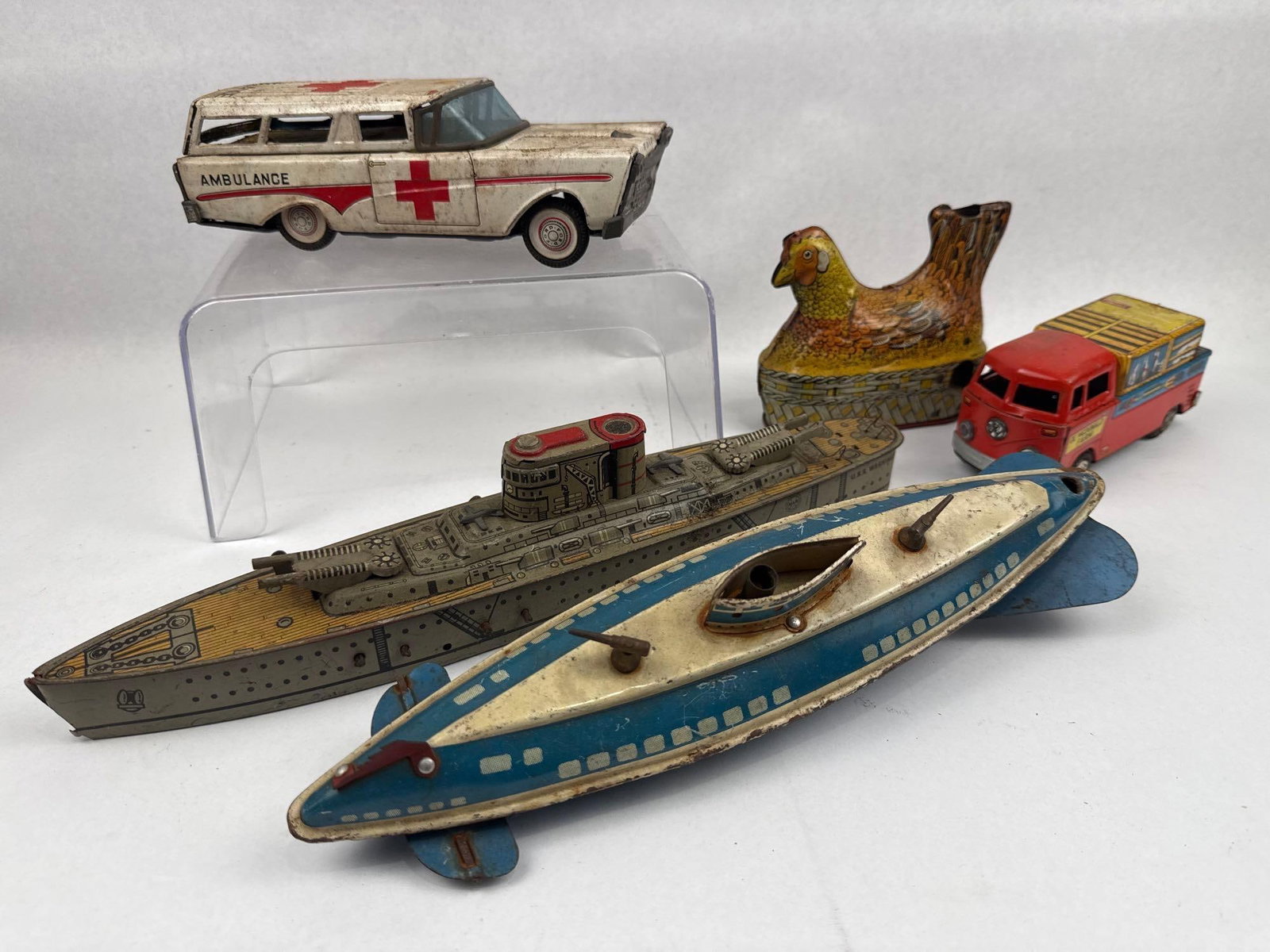 Tin-Plate Mechanical & Friction Toy: A group of mid-20th-century lithographed tin-plate toys. The collection includes a white station wagon ambulance with red cross emblems and a red stripe along the body. The hood is printed with the wo