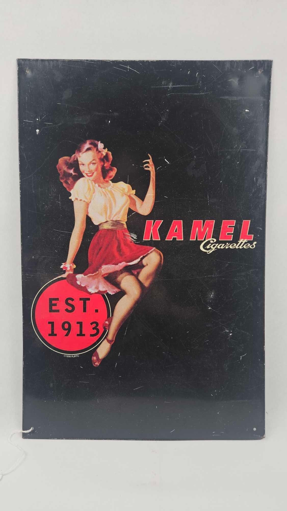 Kamel Cigarettes Advertising Metal Sign: A rectangular metal advertising sign featuring a color graphic of a mid-century style pin-up woman dressed in a white peasant blouse and red skirt. The composition is set against a black background