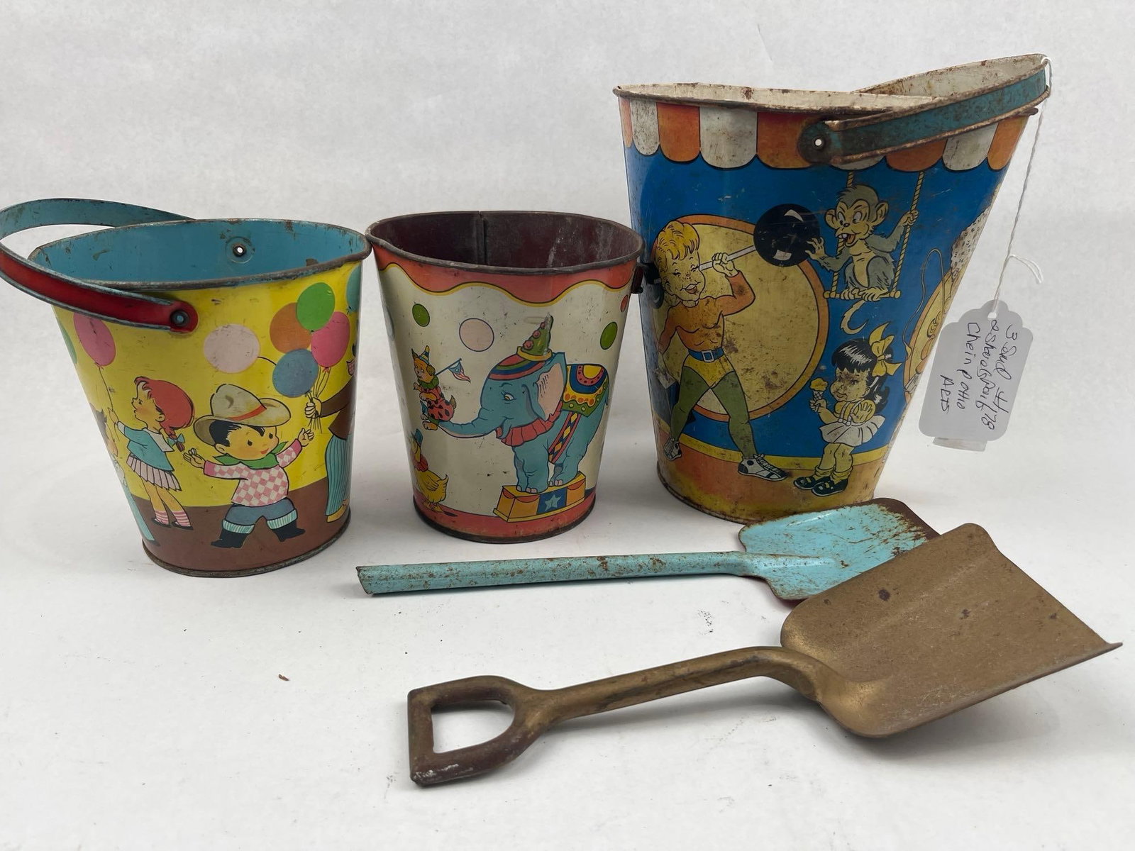 J. Chein & Ohio Art Circus Sand Pails: A collection of mid-20th century American tin-lithograph sand toys featuring circus and carnival motifs. The group includes pails of varying sizes and two metal shovels. The largest pail depicts color