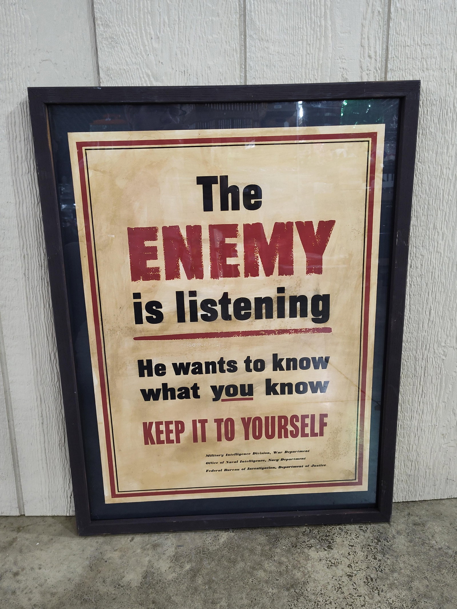 Enemy Is Listening Propaganda Poster: A framed propaganda-style poster printed on paper featuring a distressed and stained finish. The design utilizes bold black and dark red block typography against a mottled tan background. The primary