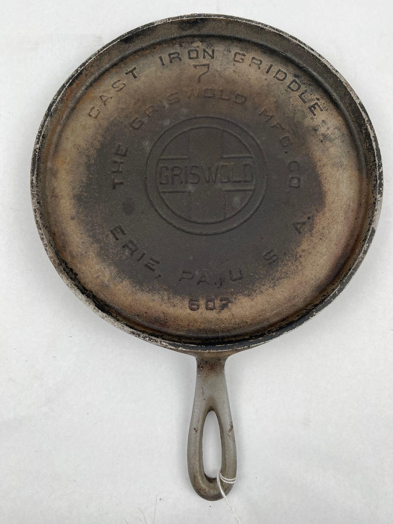 Griswold No. 7 Plated Cast Iron Griddle: A circular cast iron griddle featuring a metallic plated finish, likely nickel or chrome. The piece is designed with a low raised rim and a tapered handle with a teardrop-shaped hanging hole. The